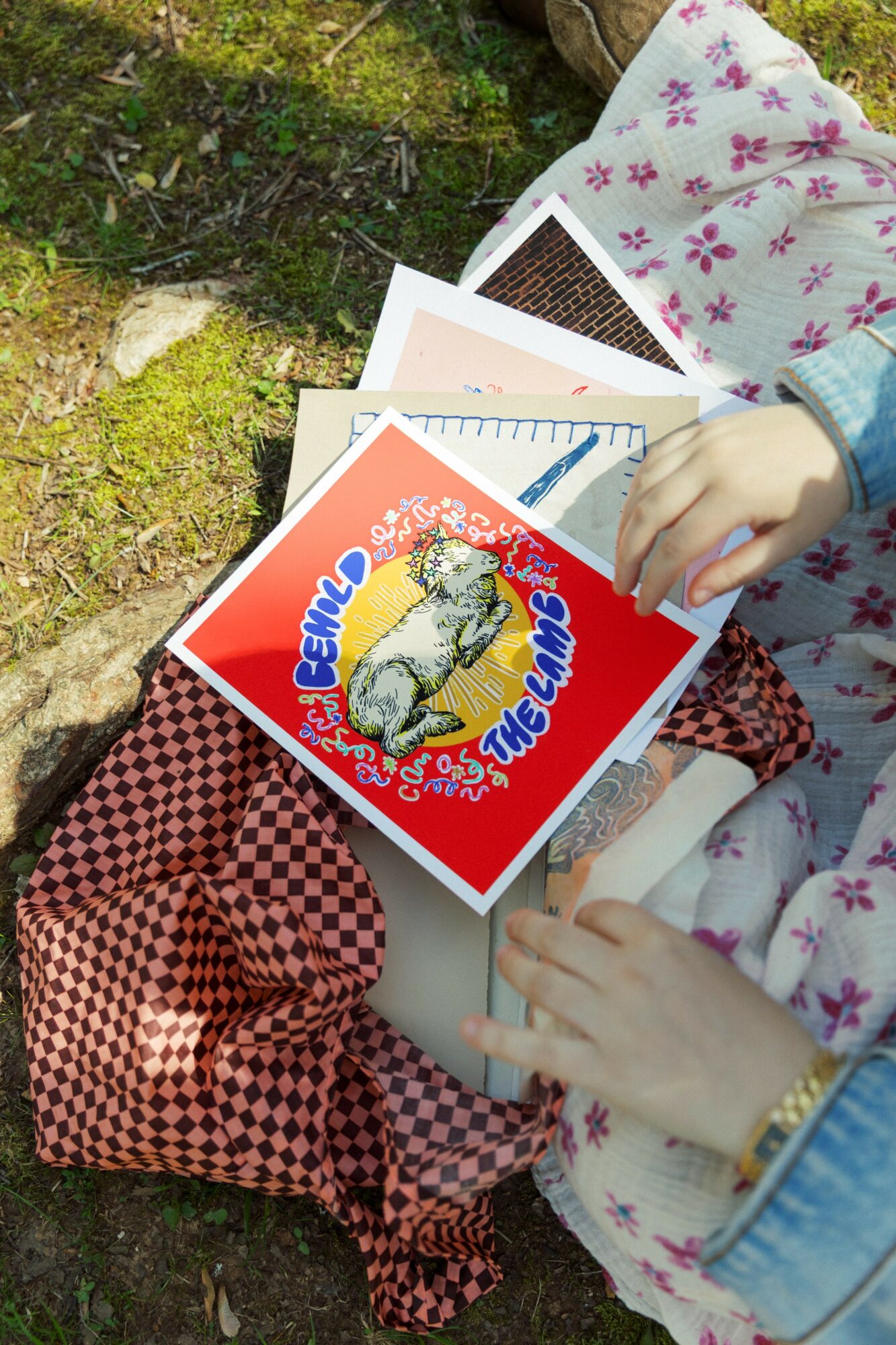 Child's hands holding a book with a colorful cover, sitting outdoors on a table with other books and a patterned cloth.