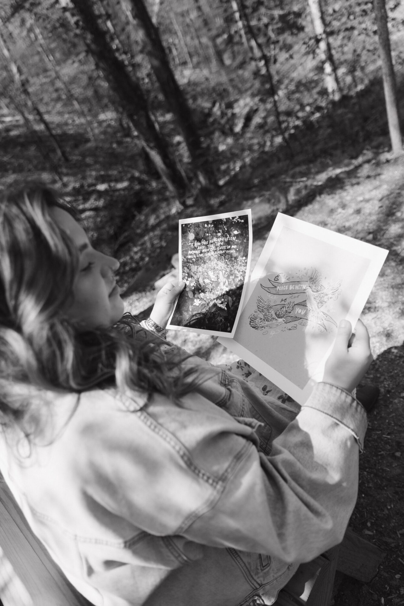 Person sitting outdoors, holding a tablet and a paper with drawings, surrounded by trees and nature.