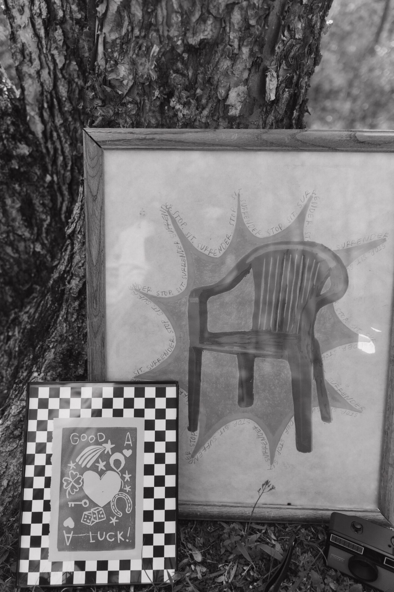 Framed drawing of a chair with a sun-like shape behind it, leaning against a tree trunk. A smaller checkered card with a cartoon character is in front.