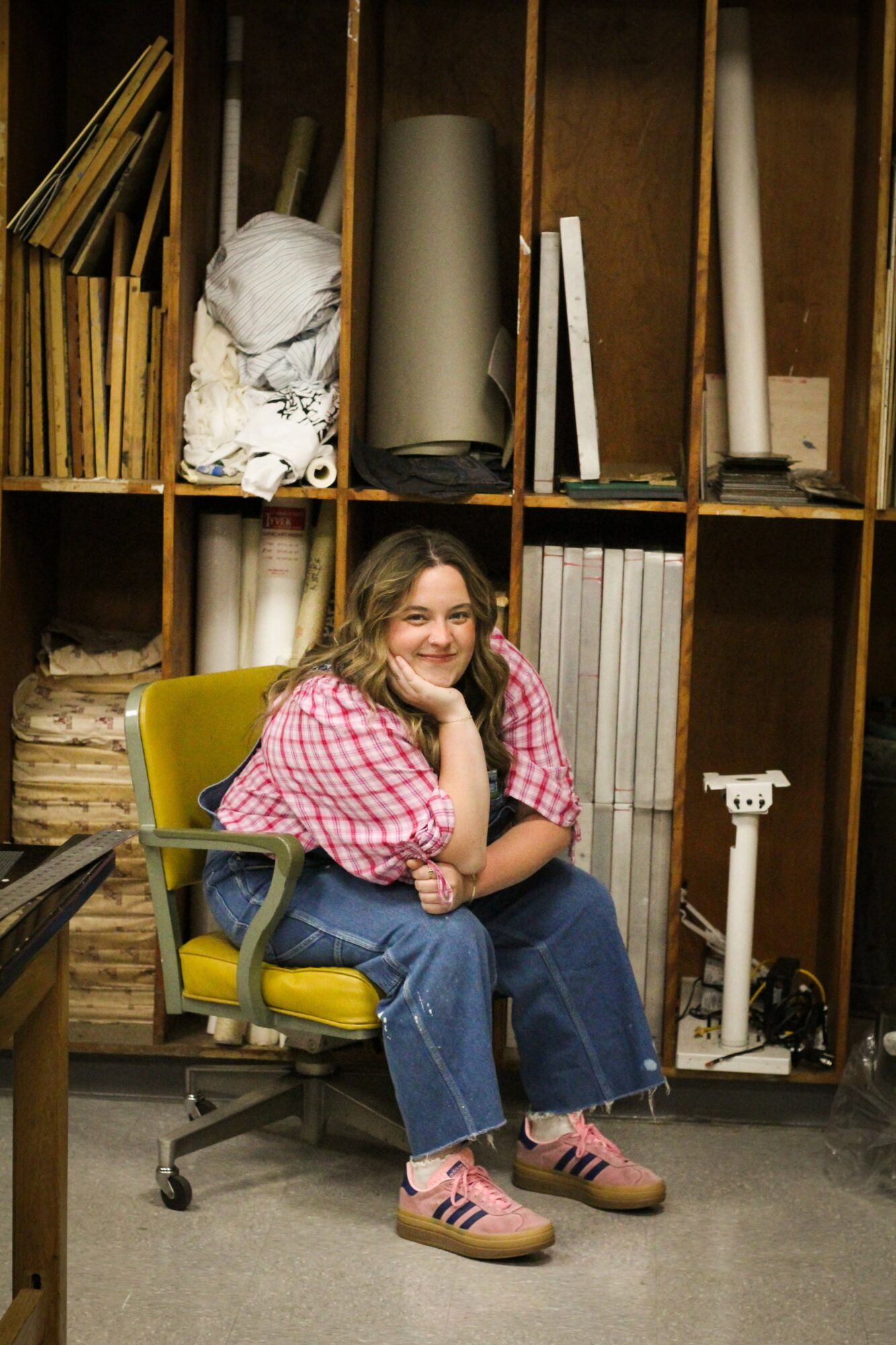 Young girl with glasses and pink checkered shirt sitting on a yellow office chair, smiling, in front of shelves with art supplies.