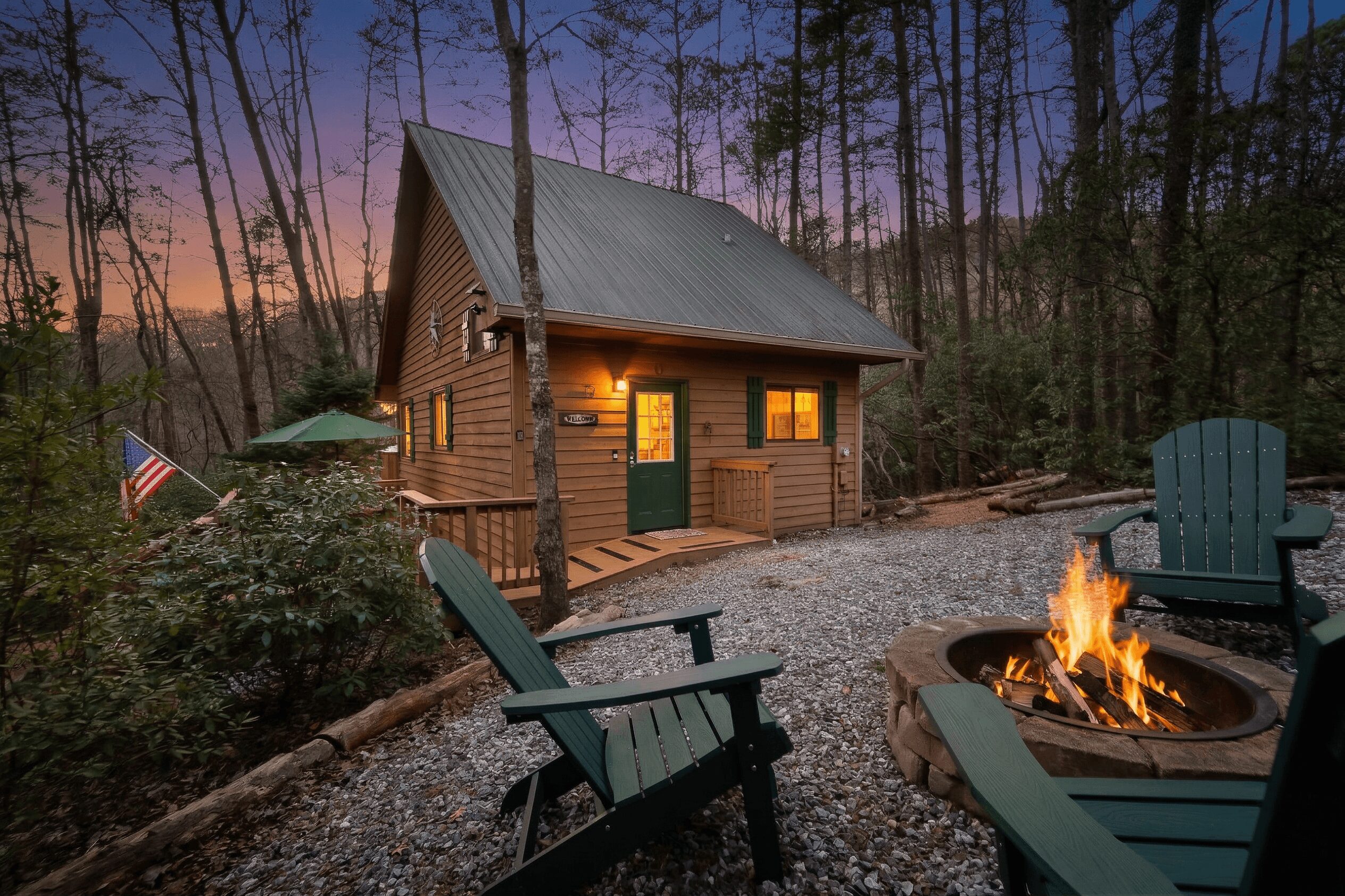 Wooden cabin with lit windows in a wooded area at sunset, surrounded by outdoor chairs and a fire pit.