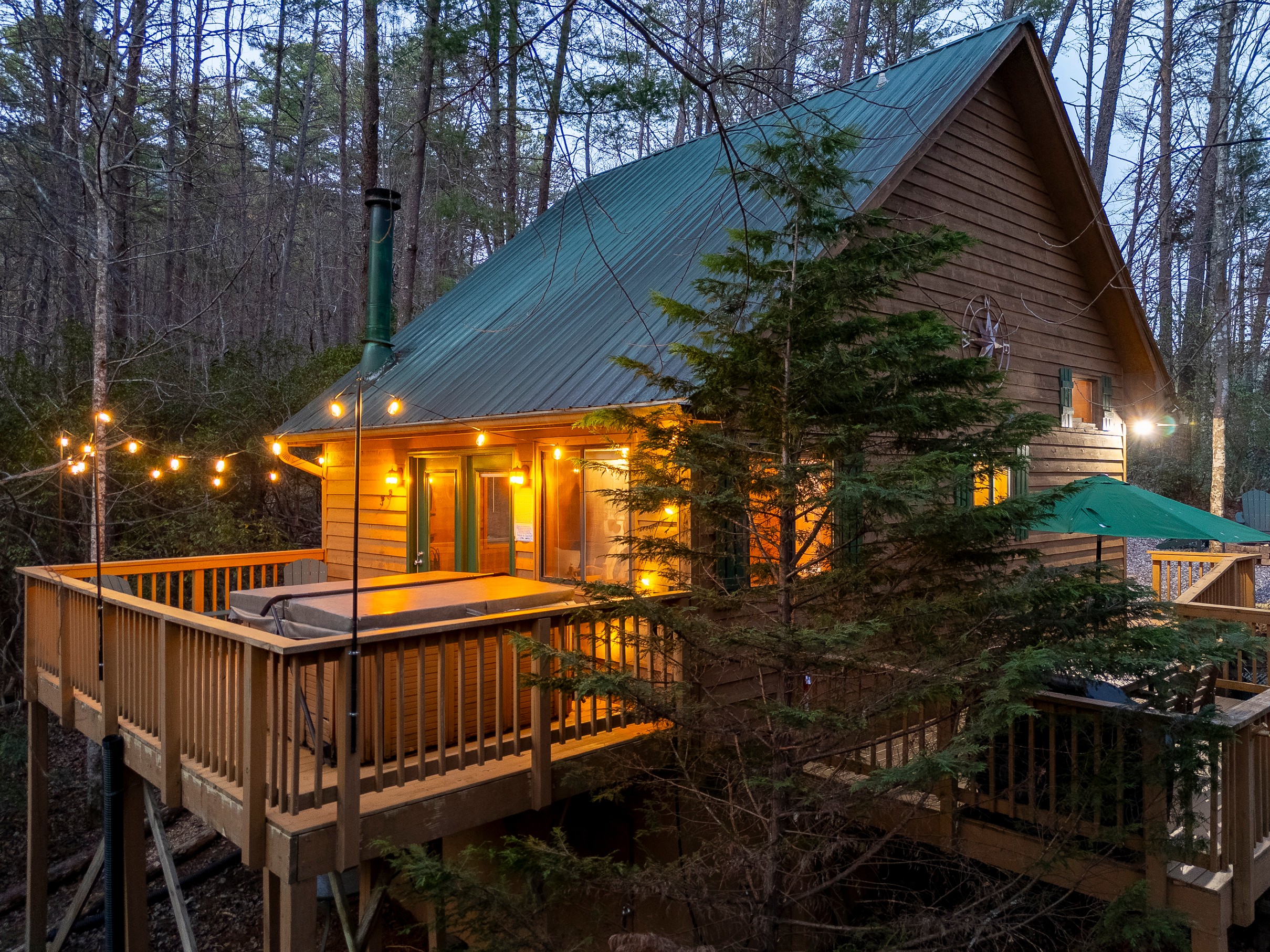 A wooden cabin with a green roof, illuminated by string lights, surrounded by trees and a deck with outdoor furniture.