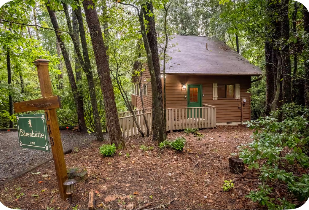 Wooden cabin in a forest with trees and a dirt path, surrounded by greenery and a signpost nearby.