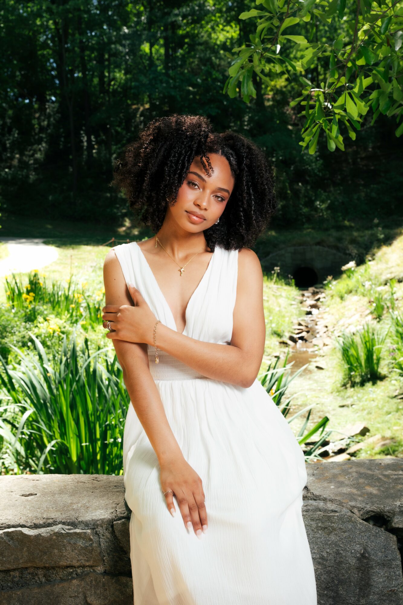 Young woman with curly hair in a white dress sitting outdoors on a stone, surrounded by greenery and trees.