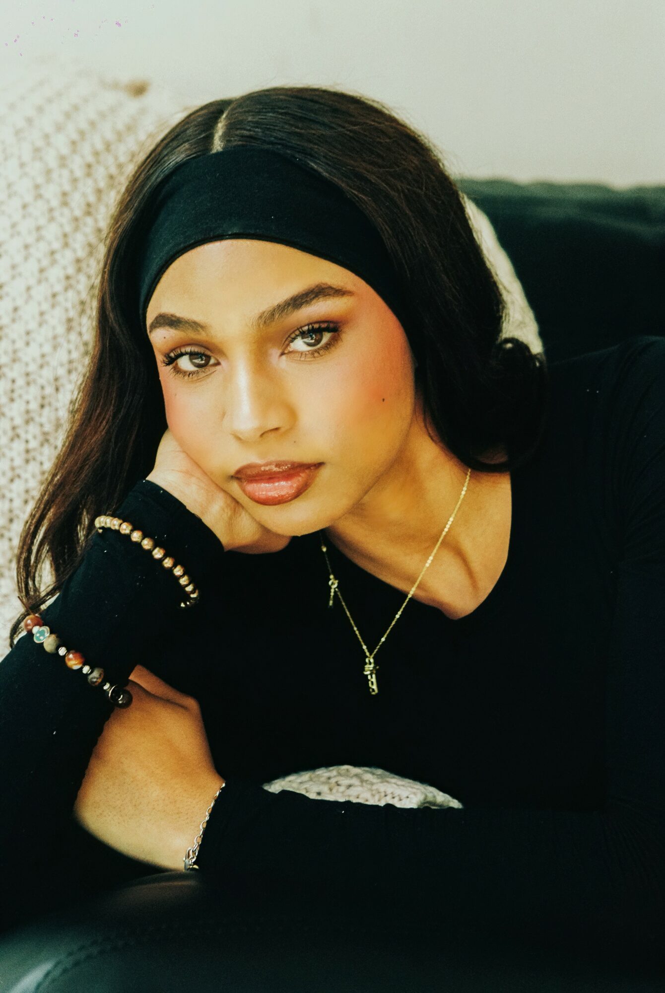 Young woman with dark hair, wearing a black headband and black top, resting her head on her hand, looking at the camera.