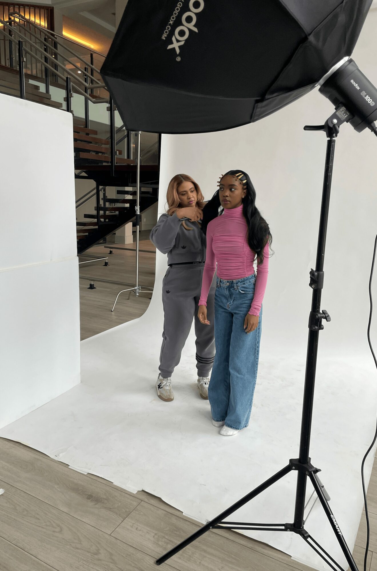 Two women stand in a photography studio with a large black umbrella light, white backdrop, and wooden flooring.