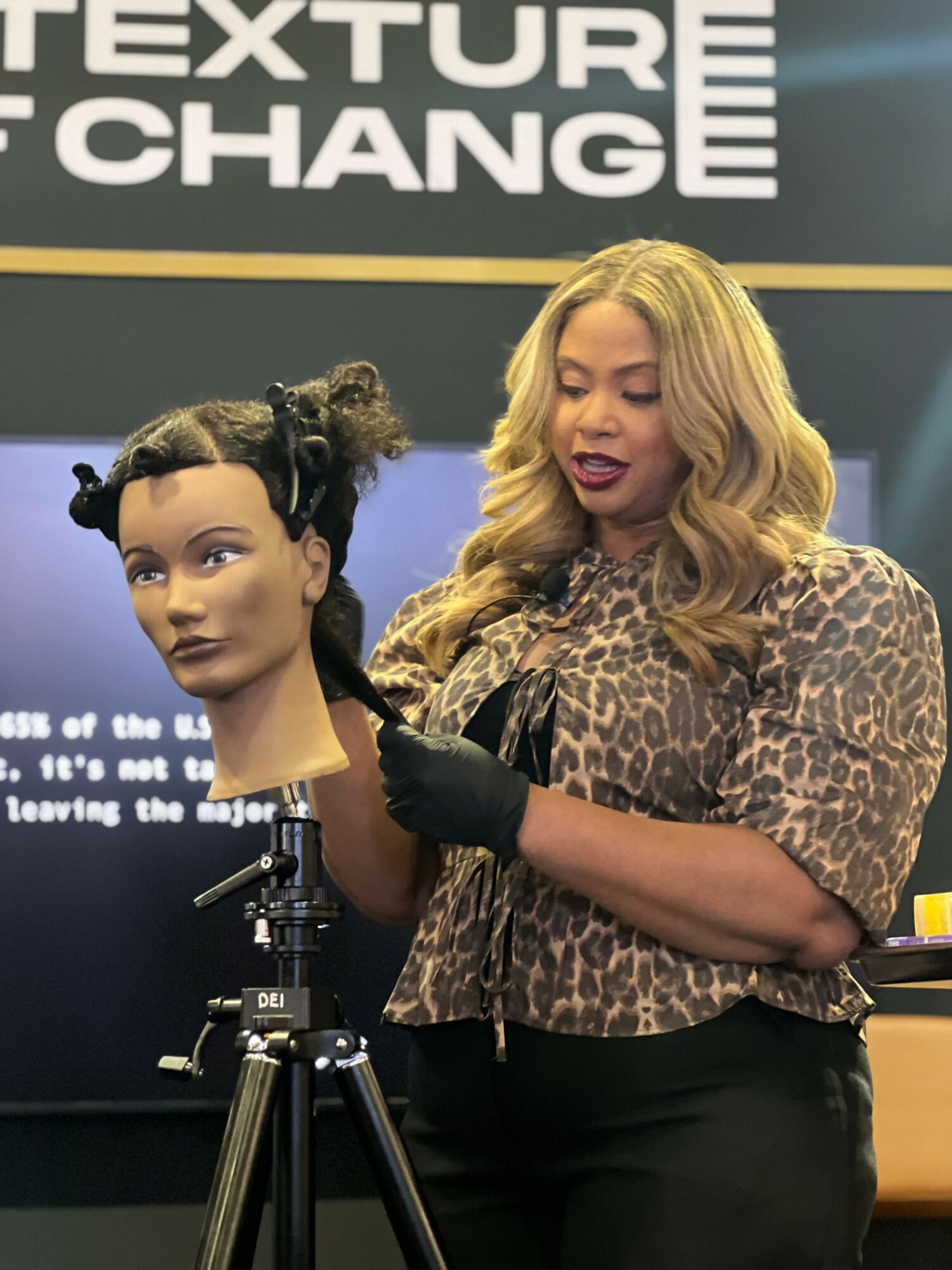 Woman with long blonde hair demonstrating hair styling on mannequin head, in front of a black background with text.