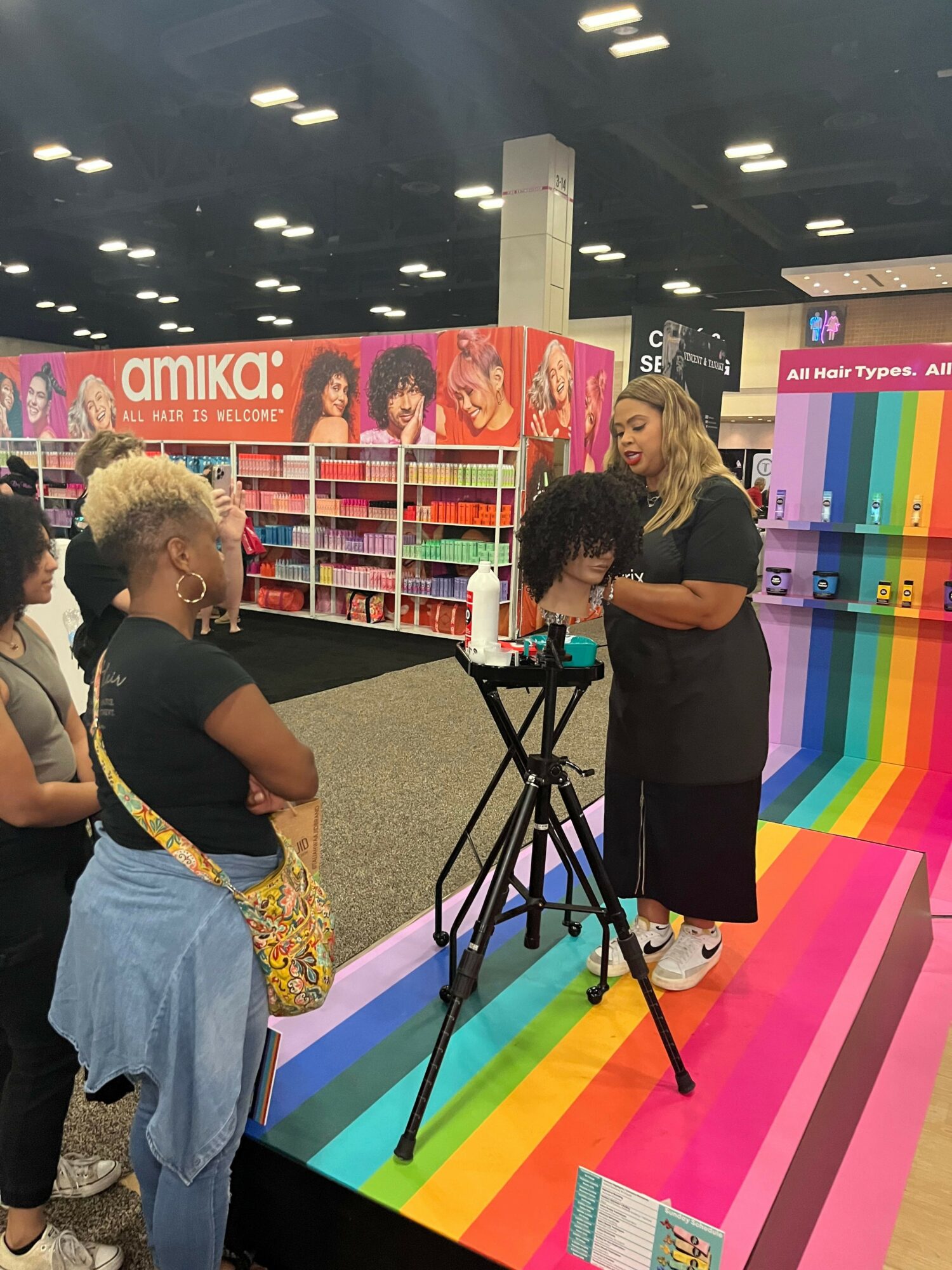 Two women browsing a colorful display of books at a store, with a rainbow-colored floor and black ceiling lights.