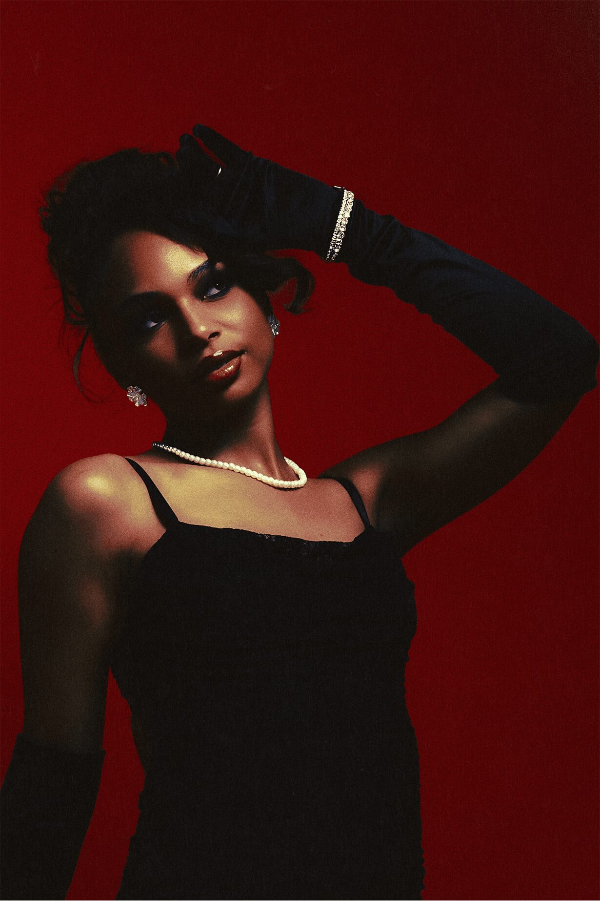 Woman in black dress with jewelry, posing against a red background, with her hand on her head.
