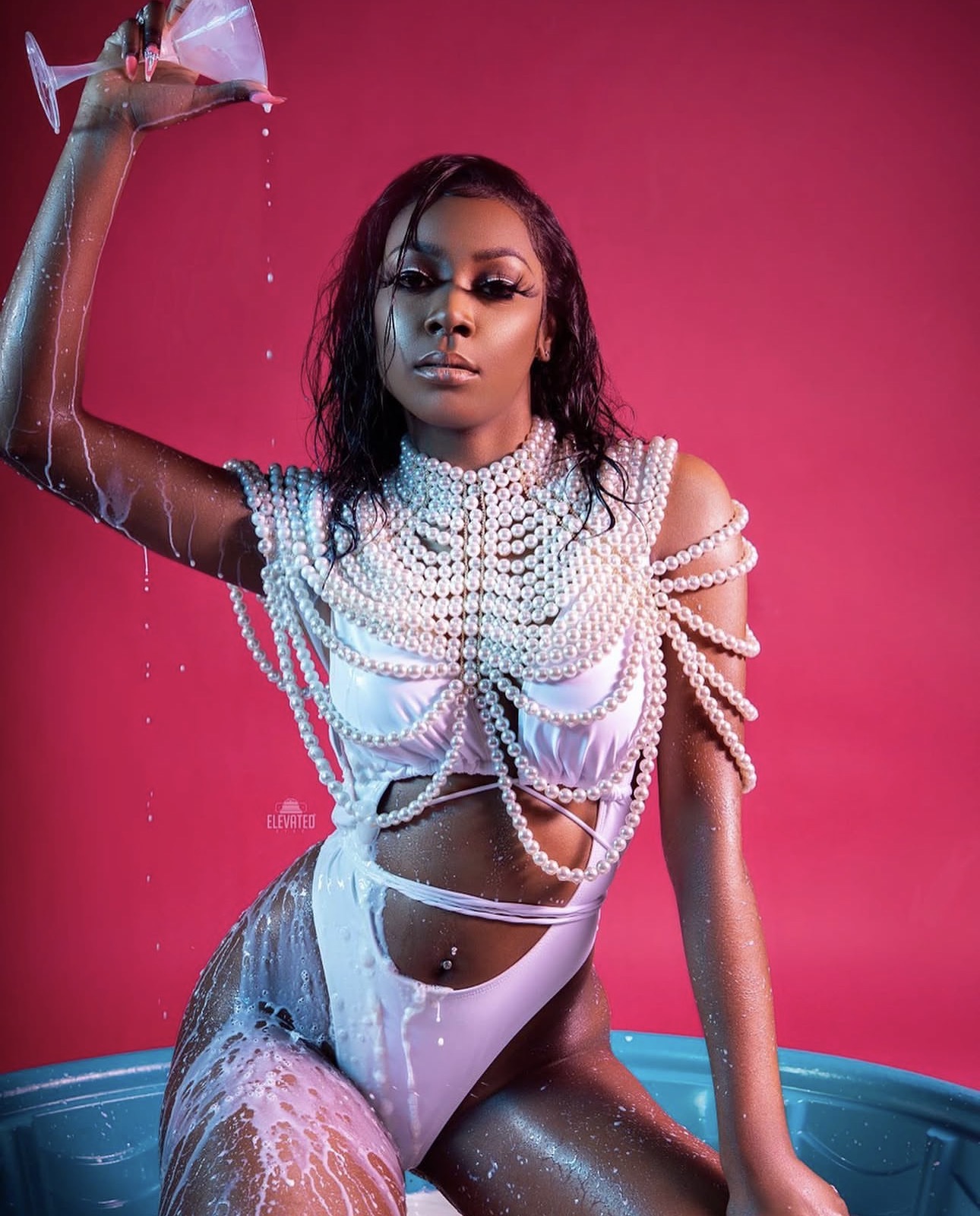 Woman in white outfit with pearl details, pouring water, pink background, blue tub, wet hair, serious expression.