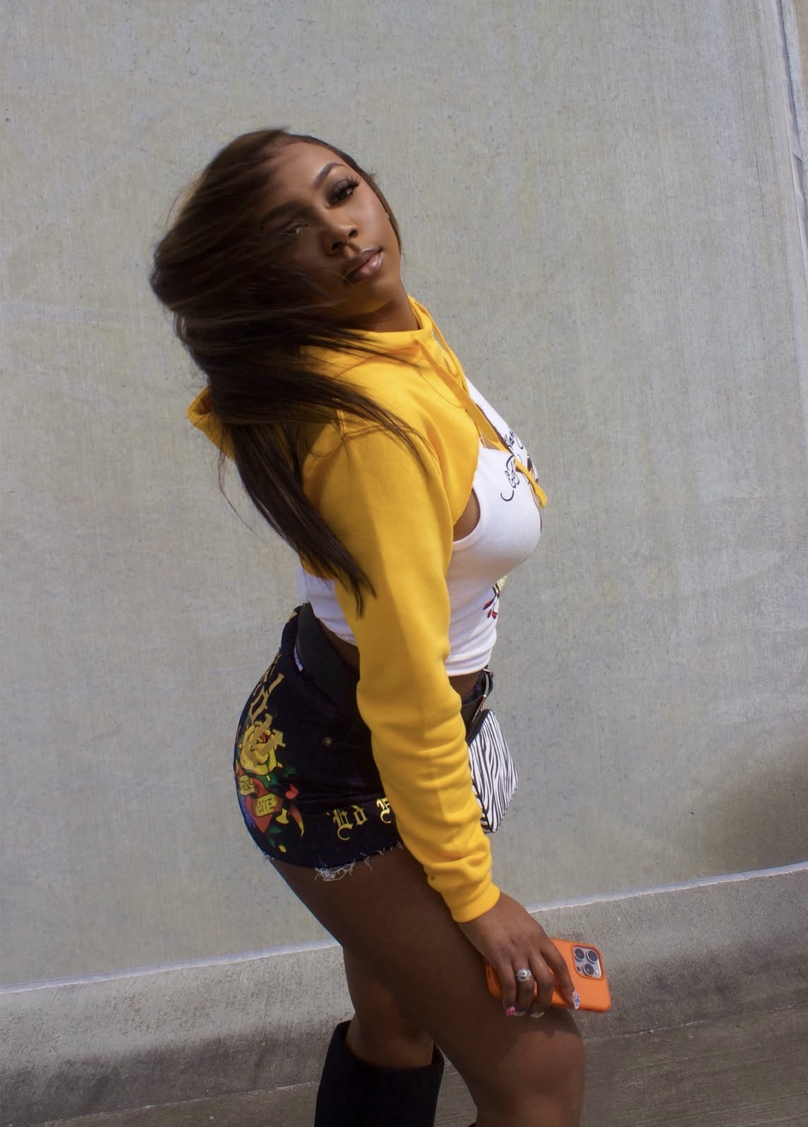 Young woman with long hair wearing a yellow hoodie and floral shorts, holding a phone, standing against a plain wall.