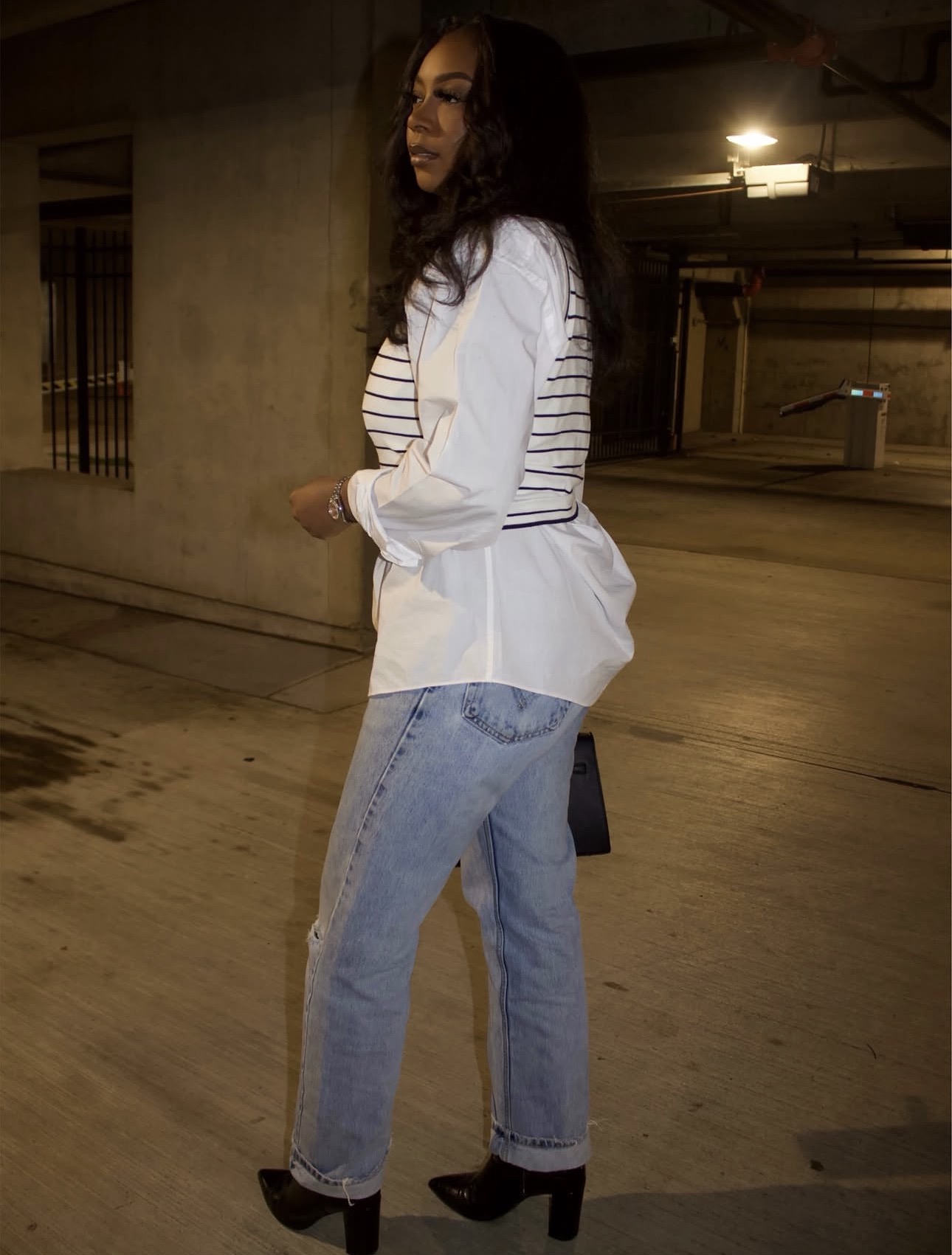 Woman standing in a parking garage, wearing a striped shirt, white jacket, jeans, and high heels, looking to the side.