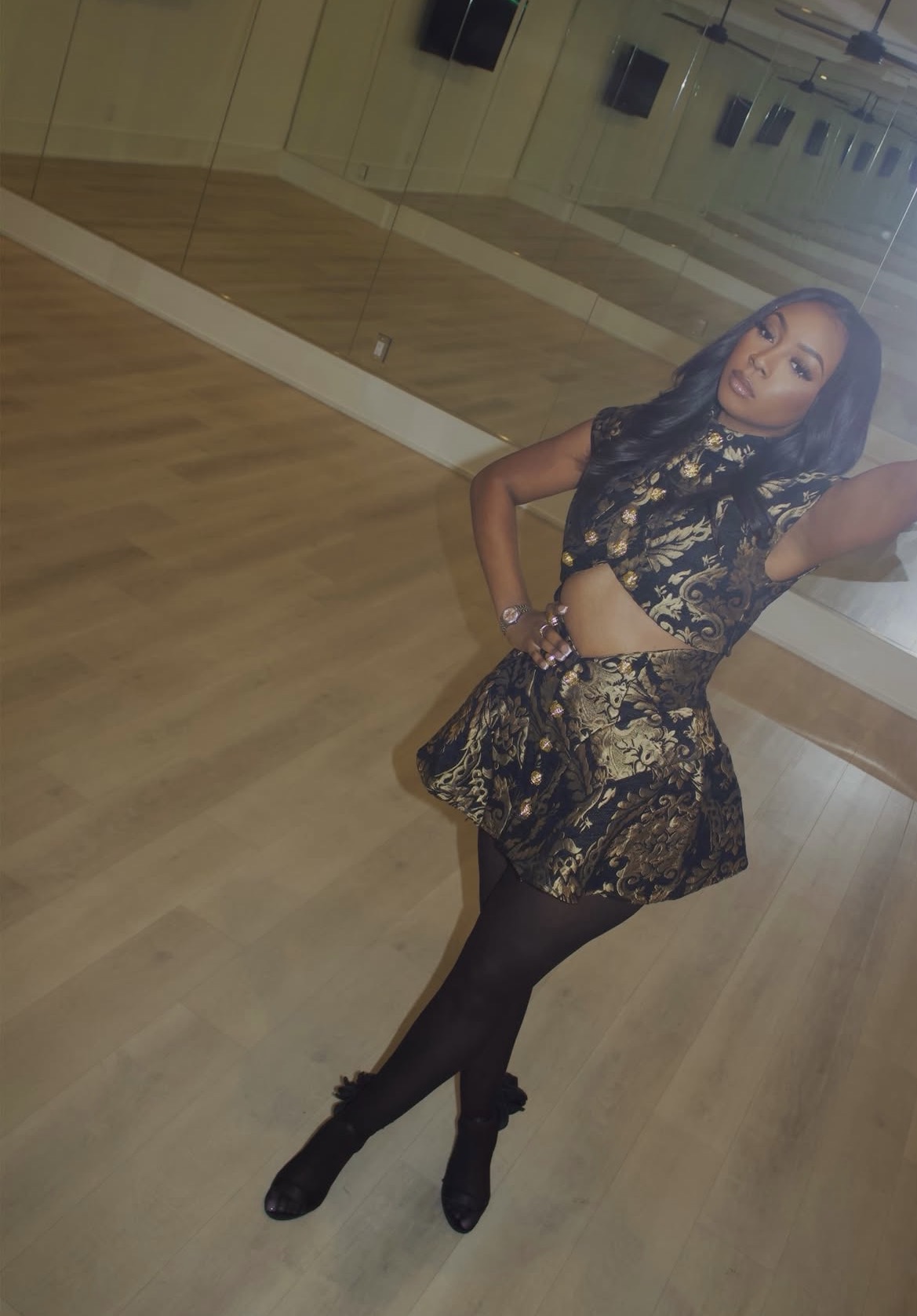 Woman in a black and gold dress taking a selfie in a dance studio with wooden floors and mirrors.