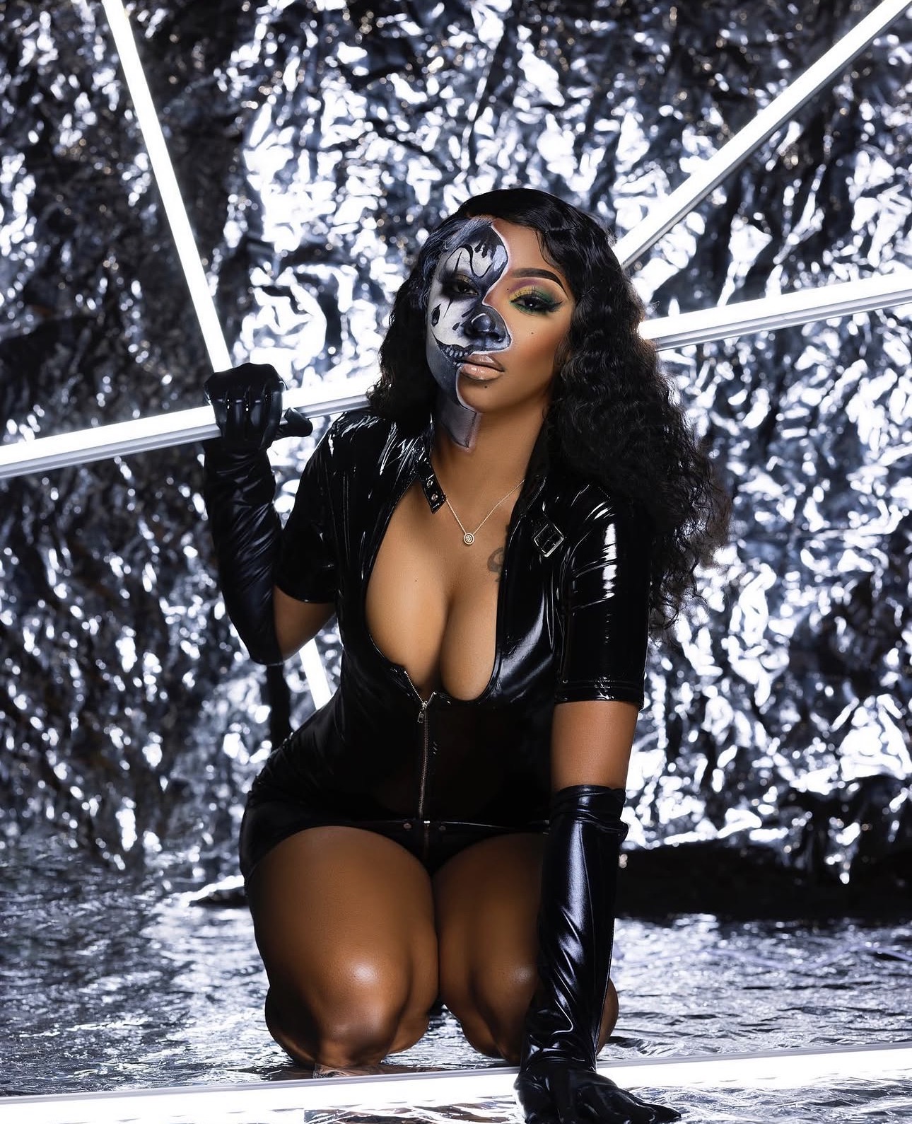 Woman with black curly hair in a black shiny outfit, holding white light tubes, kneeling in front of a metallic background.
