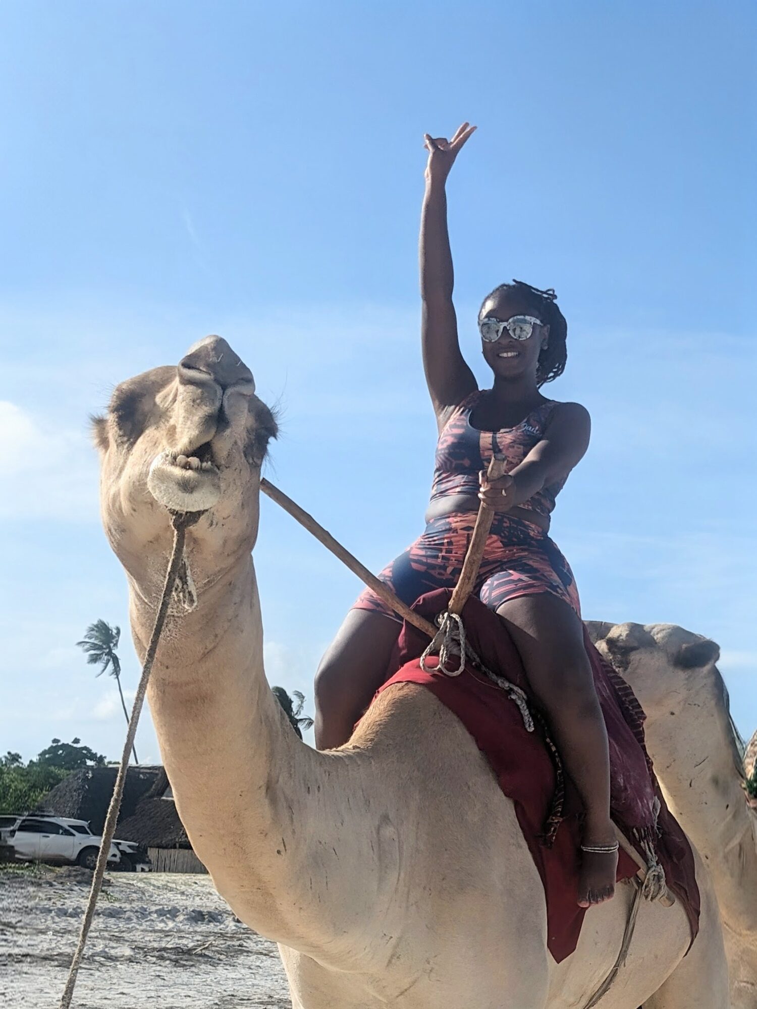Woman riding a camel, raising one arm, smiling, against a clear blue sky.