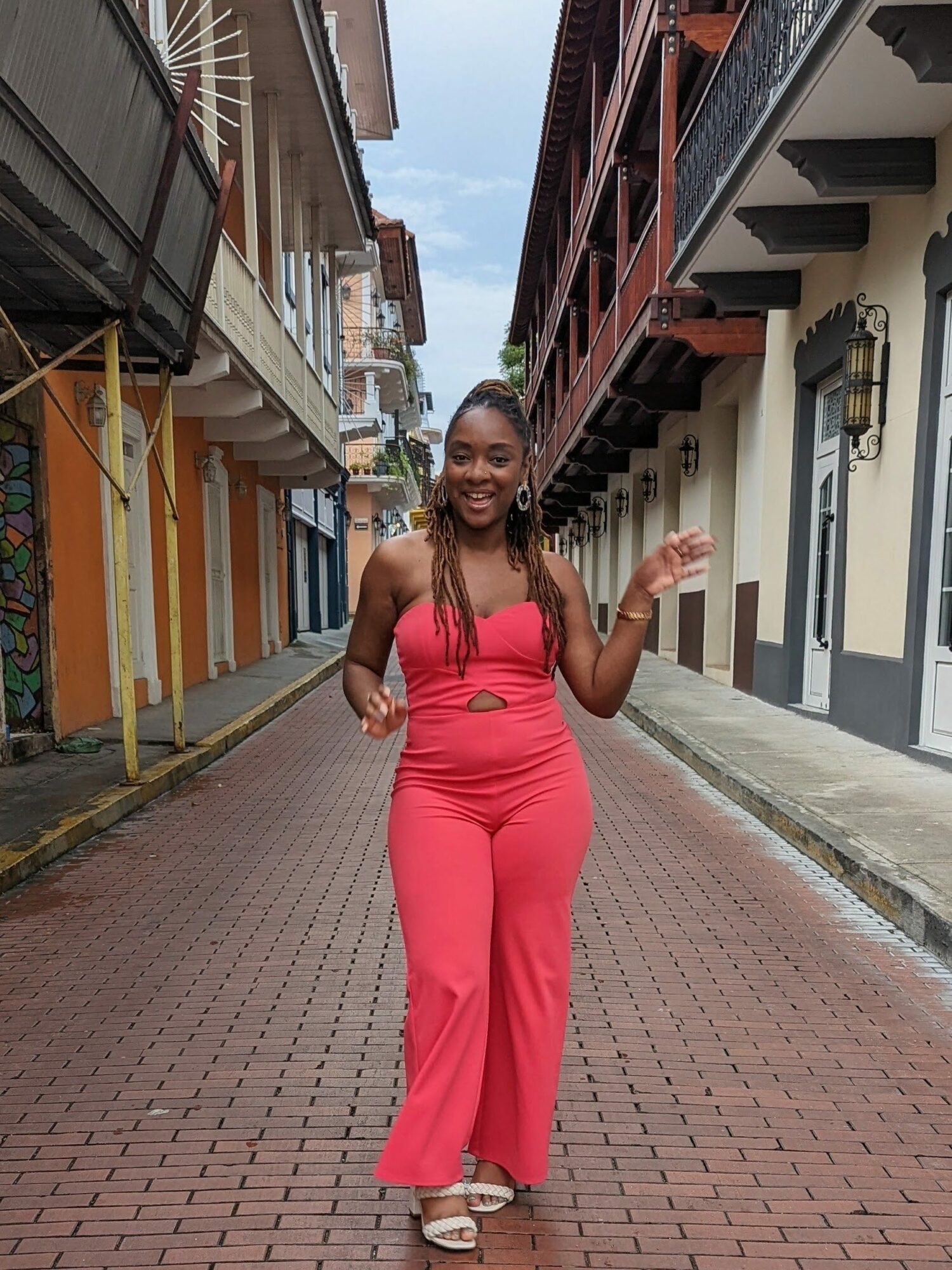 Smiling woman in pink jumpsuit walking on a colorful city street, holding her hand up, buildings on both sides.