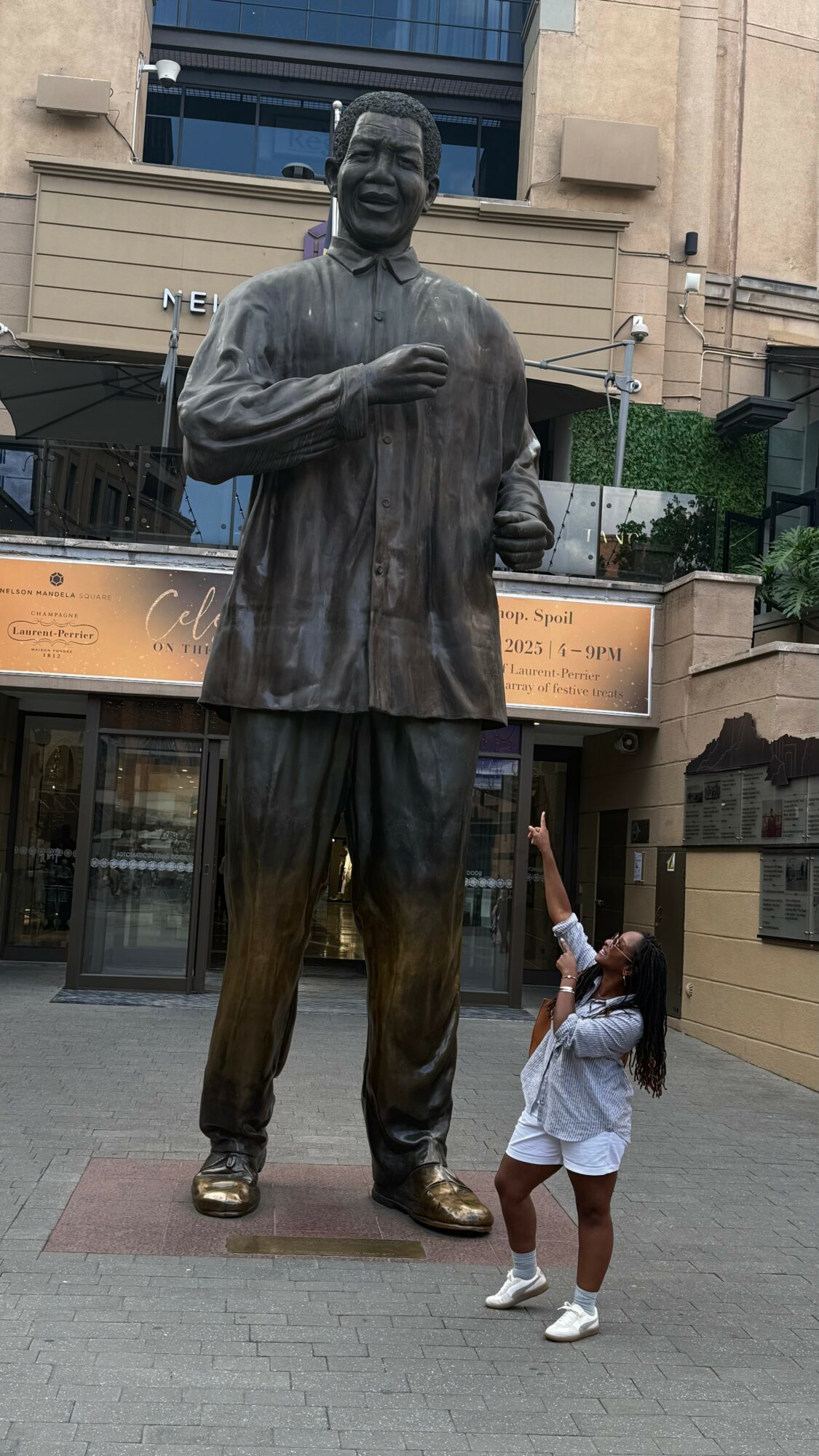 Child reaching up to a large bronze statue of a smiling man in traditional attire, standing outdoors in an urban setting.