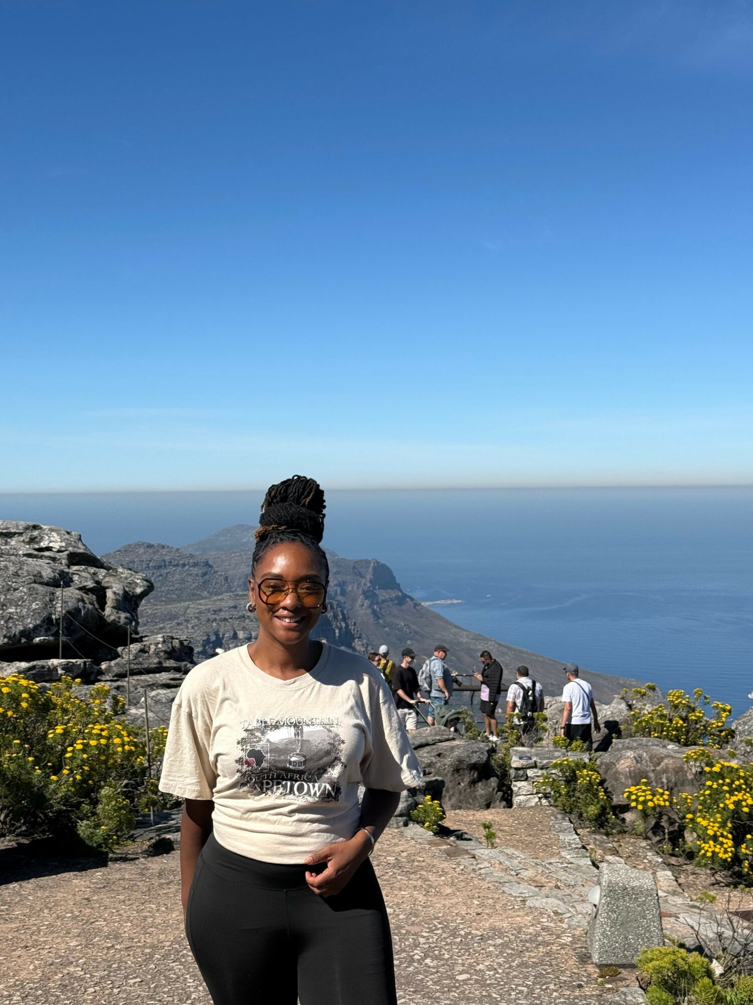 Woman standing outdoors near water and mountains, with people in the background, sunny day, wearing a white top and black pants.