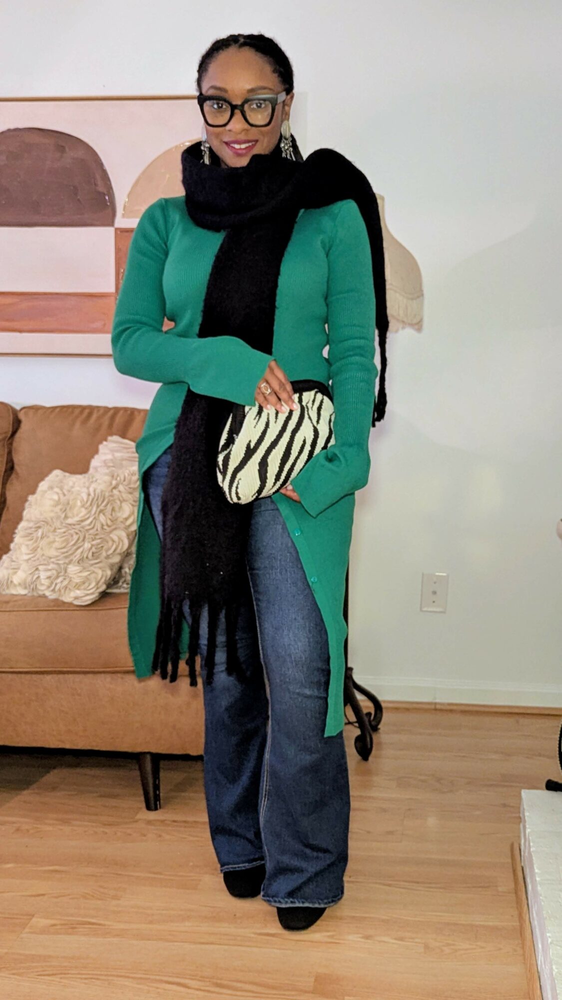 Woman standing indoors, wearing glasses, a green coat, black scarf, and holding a zebra-patterned clutch, smiling.