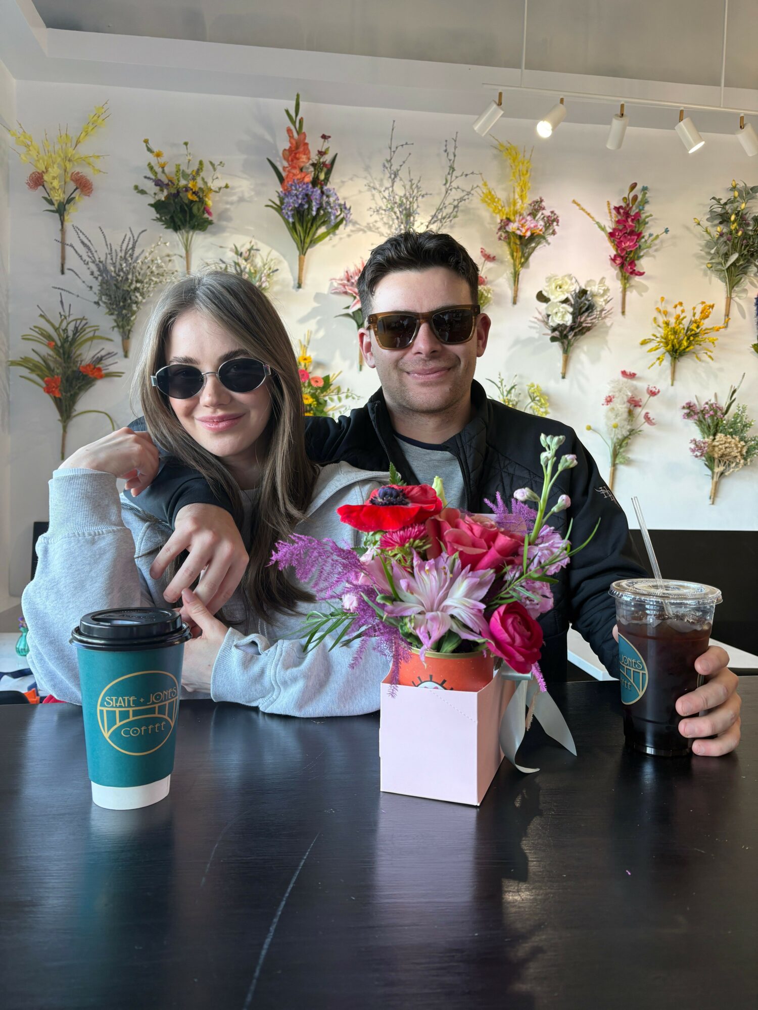 Two women with sunglasses sit at a table with a flower arrangement and drinks, smiling at the camera.