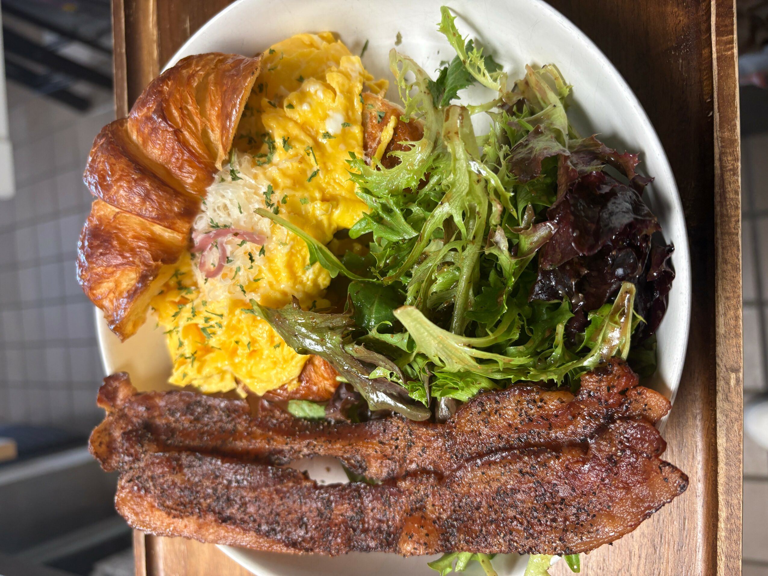Plate with scrambled eggs, croissant, mixed greens salad, and bacon strips.