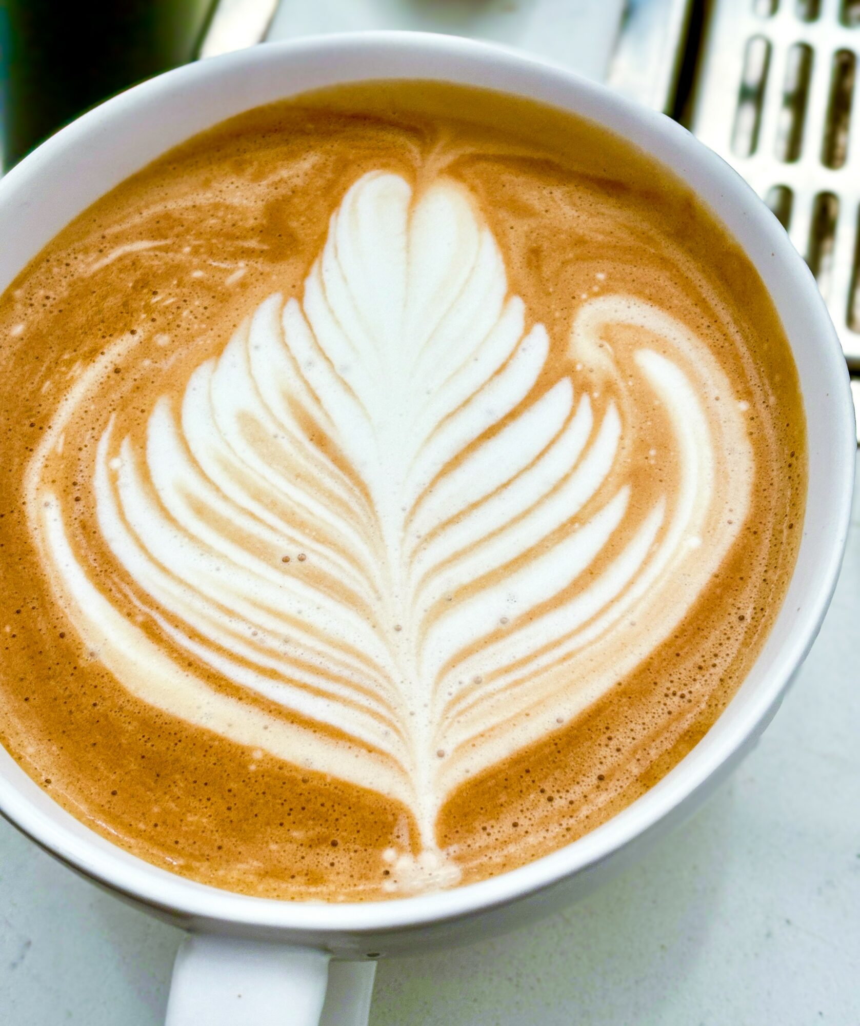 Close-up of a latte with a leaf-shaped foam design on top, in a white cup on a light surface.
