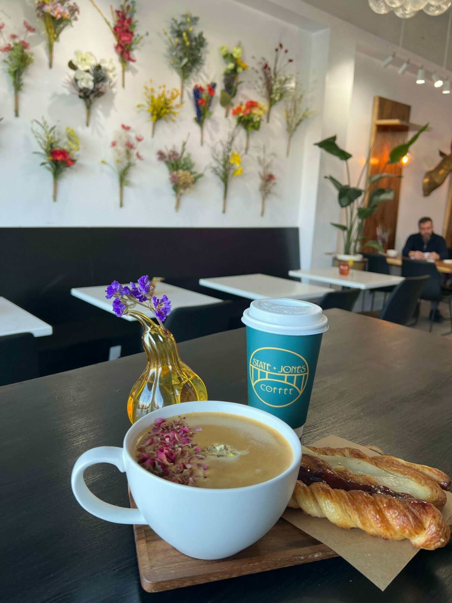 Coffee cup with pink flowers, pastry, and a drink on a table in a cafe with floral wall decor.