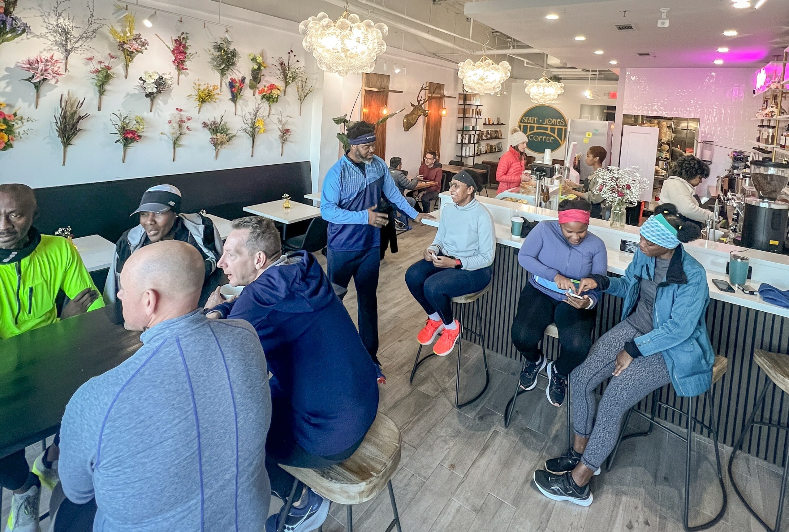 People sitting and standing in a cafe with floral wall decor and chandeliers, some using tablets or phones.
