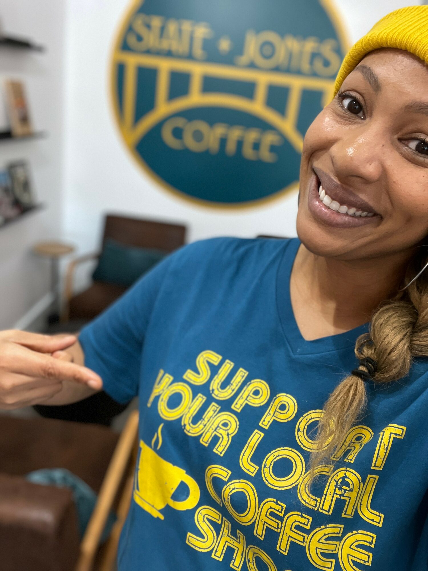 Smiling woman wearing a yellow beanie and blue shirt pointing at text on her shirt, in a coffee shop setting.