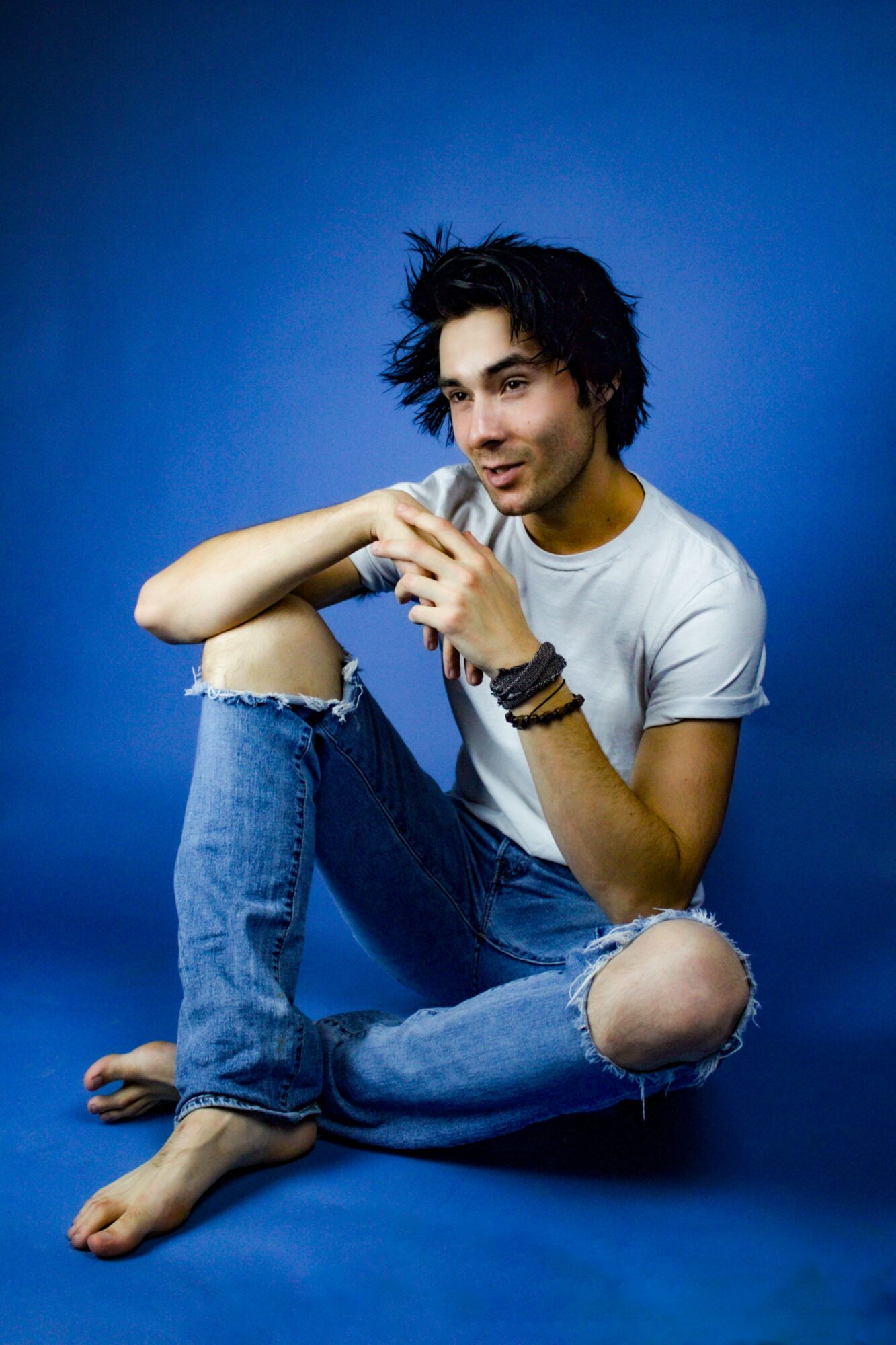 Young person with dark, curly hair sitting on the floor against a blue background, wearing a white t-shirt and ripped jeans, holding a phone.