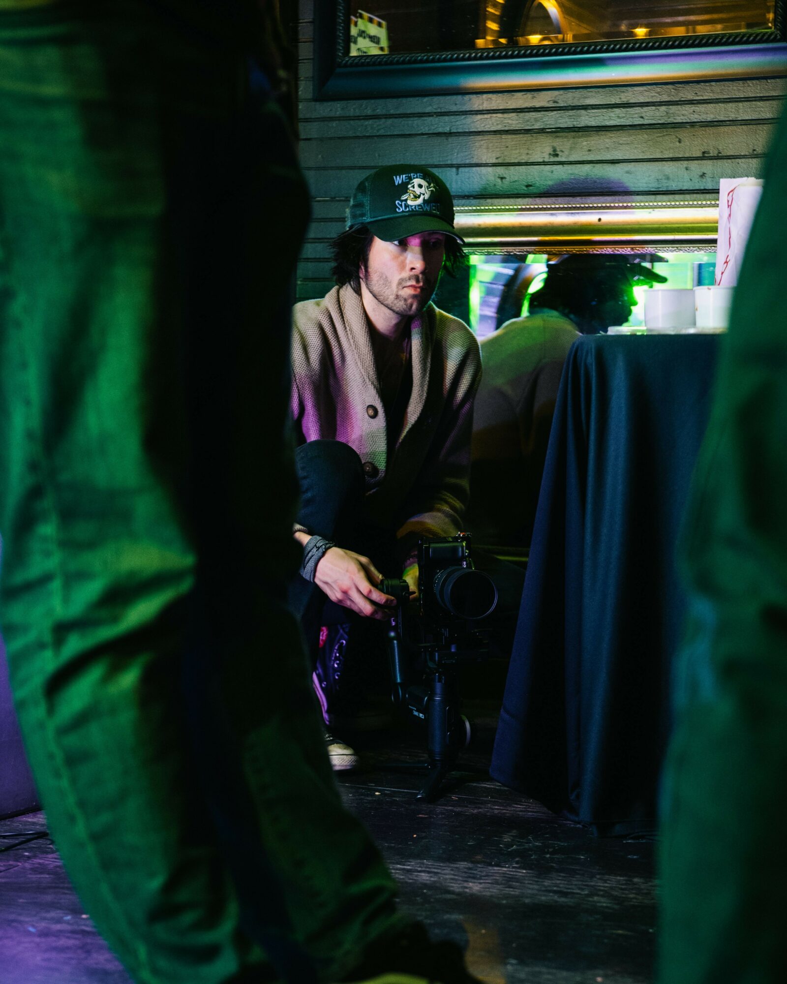 Person sitting behind a table with a camera, wearing a cap, in a dimly lit environment with colorful lighting.