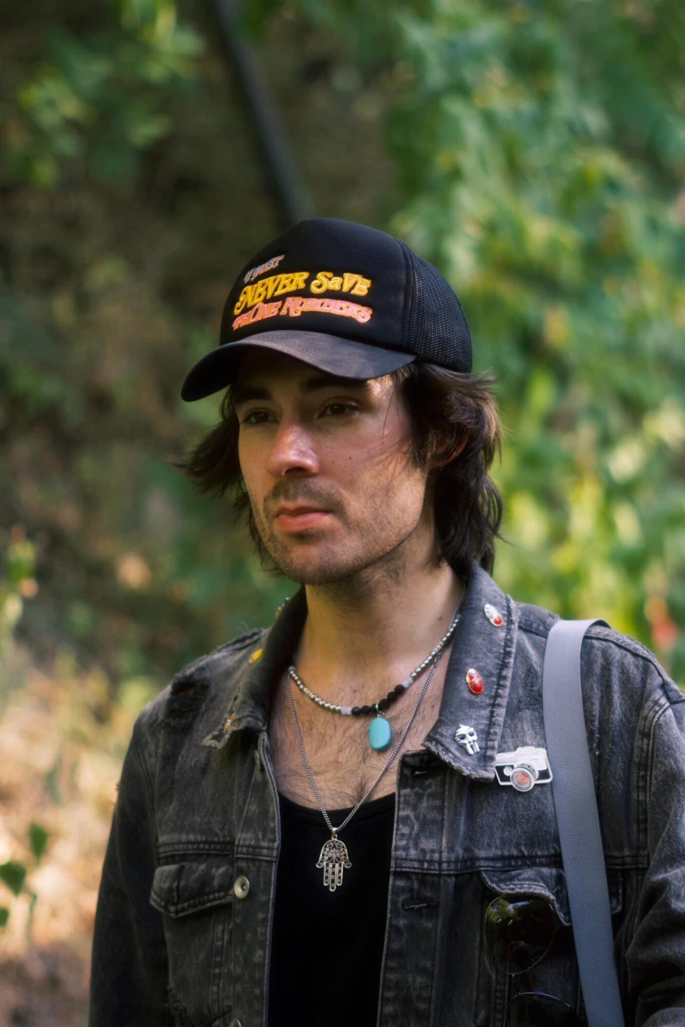 Young man with dark hair, wearing a black cap, denim jacket, and backpack outdoors with green foliage.