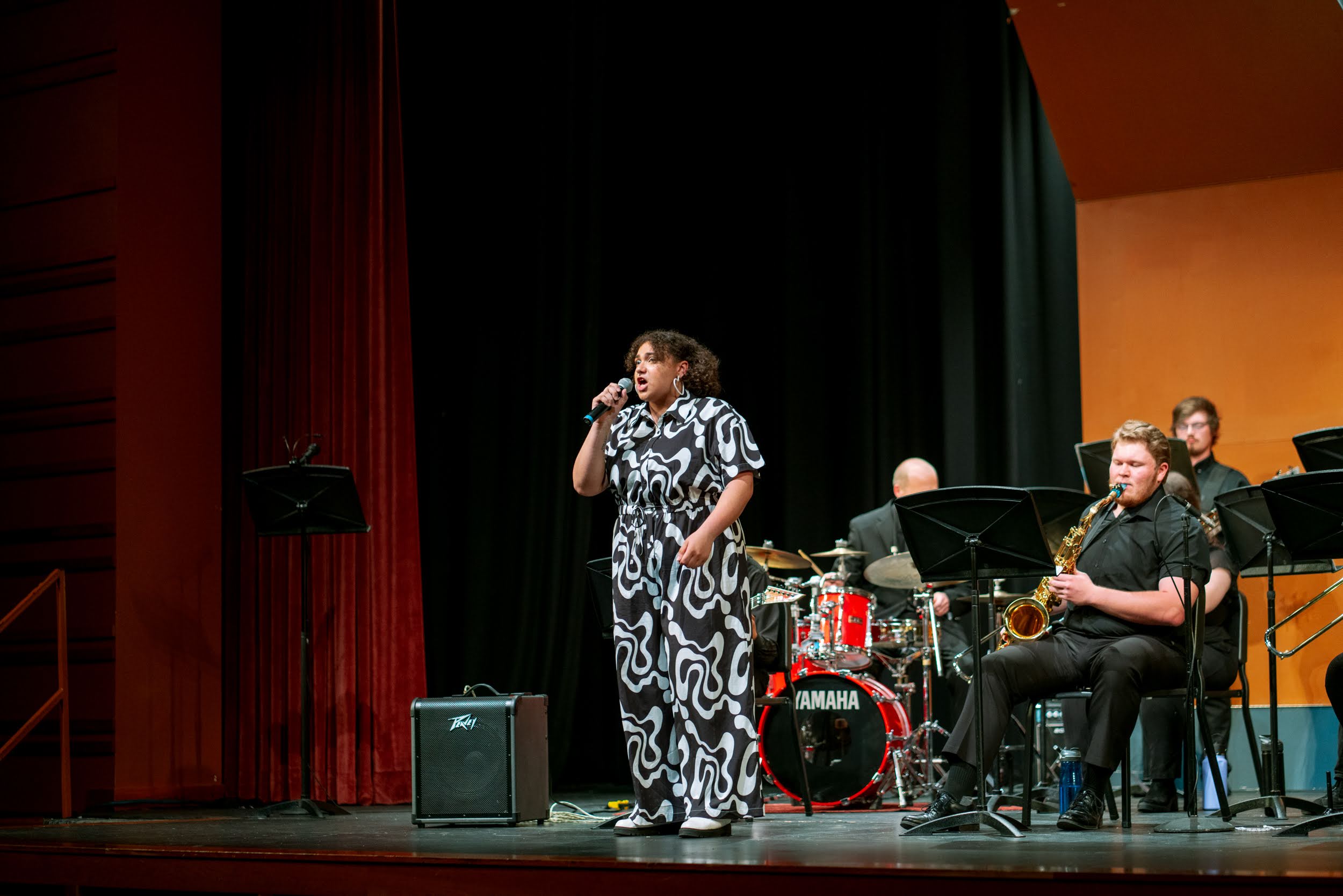 A woman sings into a microphone on stage with musicians, drums, and a keyboard, in front of a black curtain.