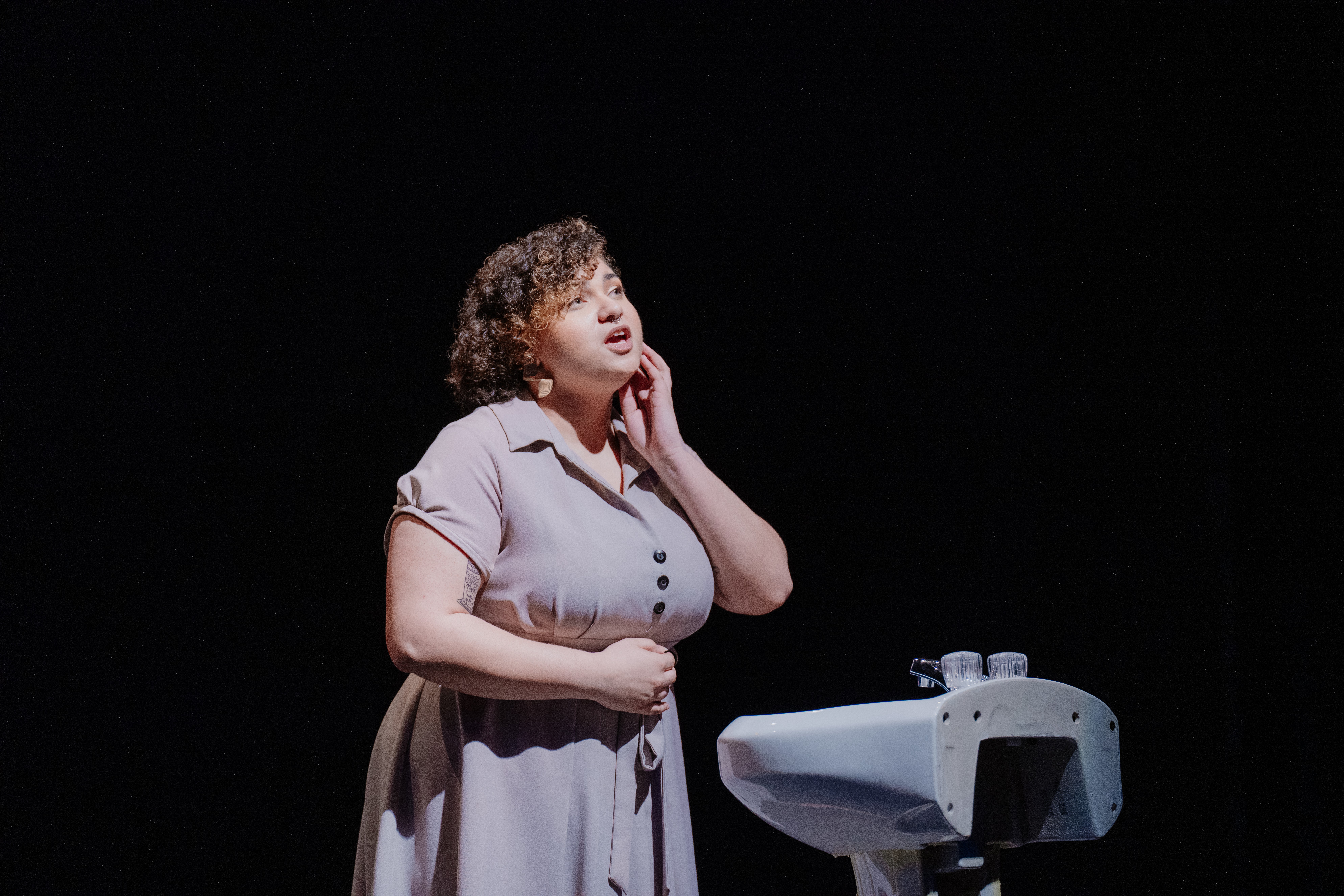 Woman with curly hair touching her neck, standing near a sink, against a black background.