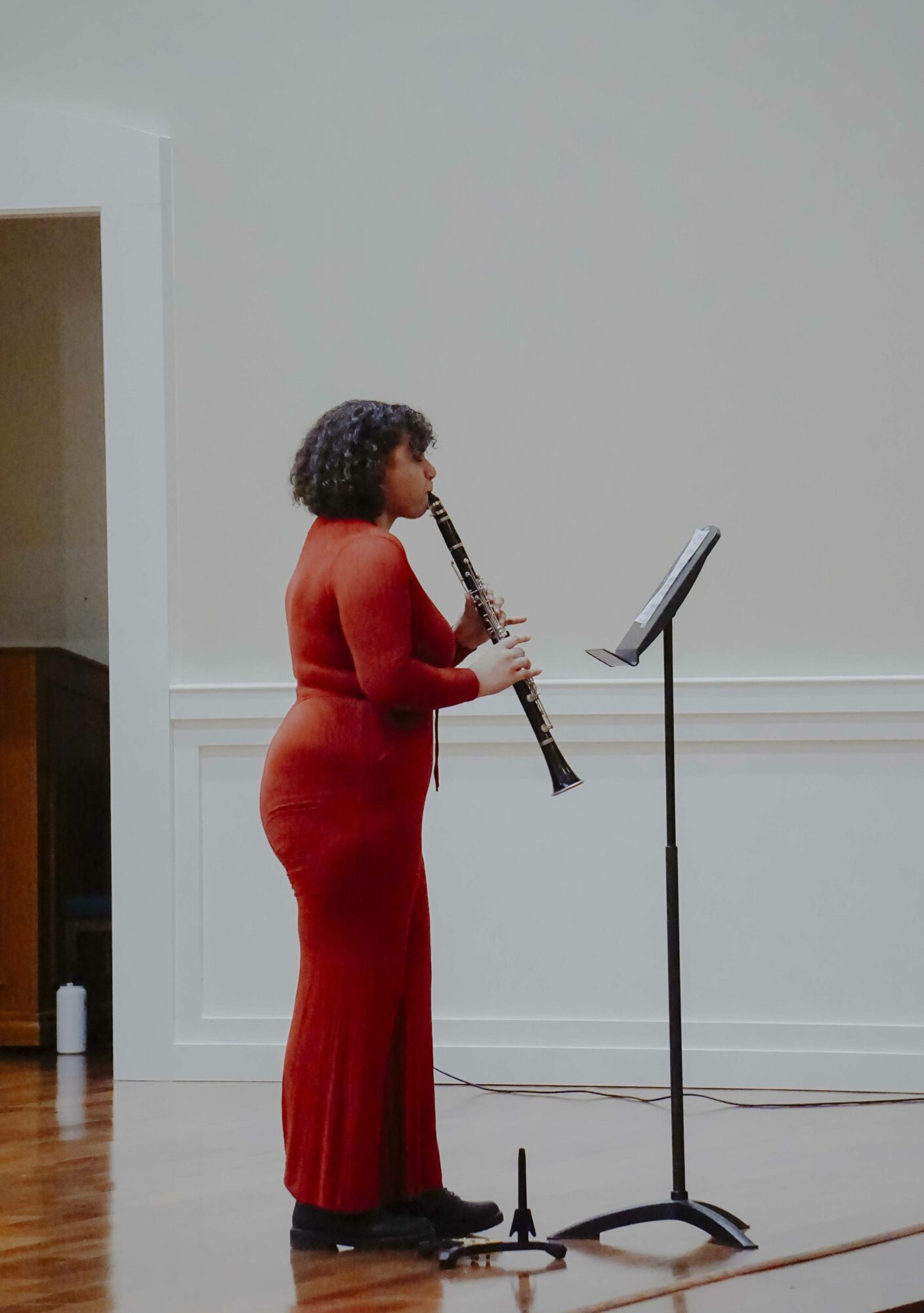 Woman in red dress playing a clarinet on stage with music stand, plain background, wooden floor.