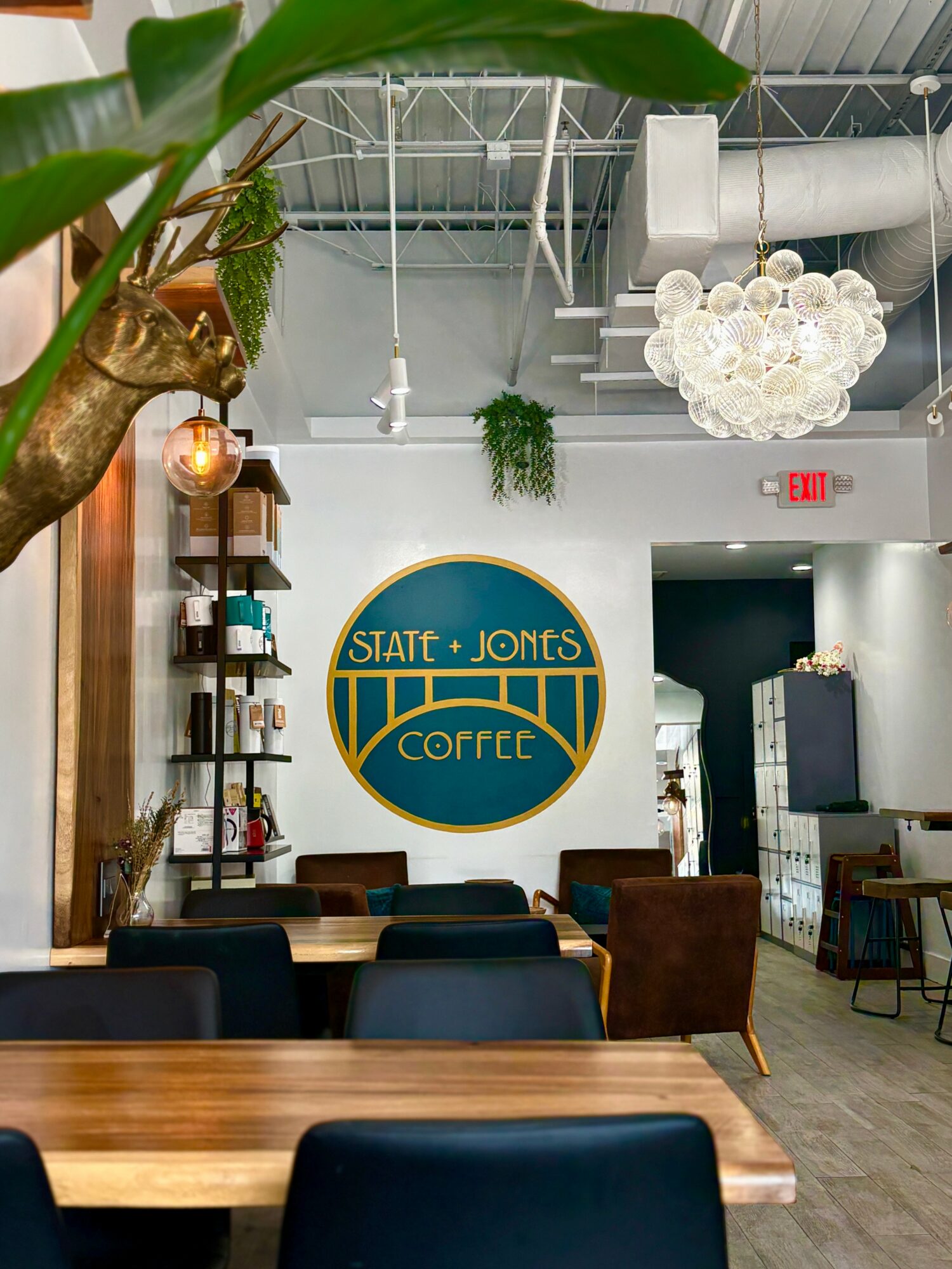 Interior of a coffee shop with a large circular sign reading 'State + Jonts Coffee' on the wall, tables, chairs, and a chandelier.