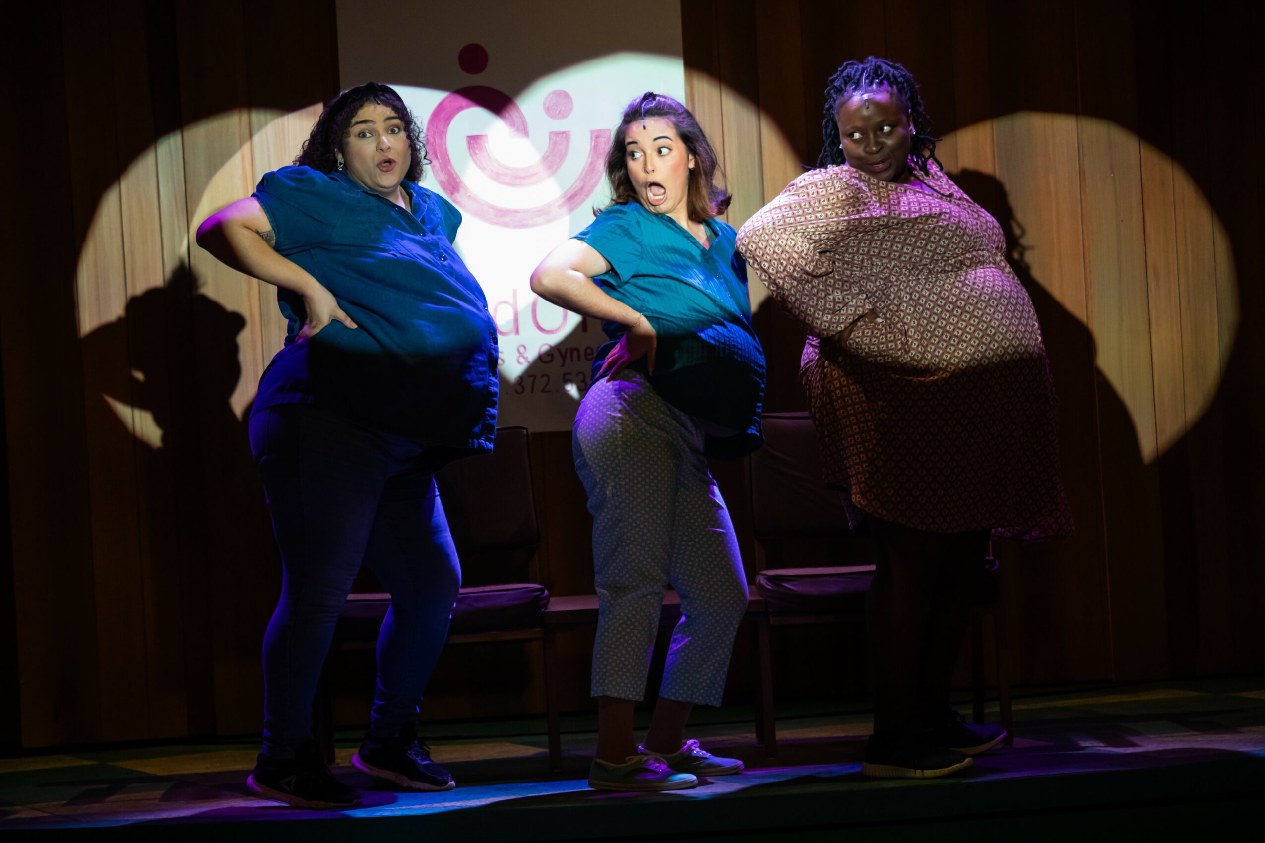 Three women stand on stage with spotlights, posing confidently with hands on hips, in front of a curtain backdrop.