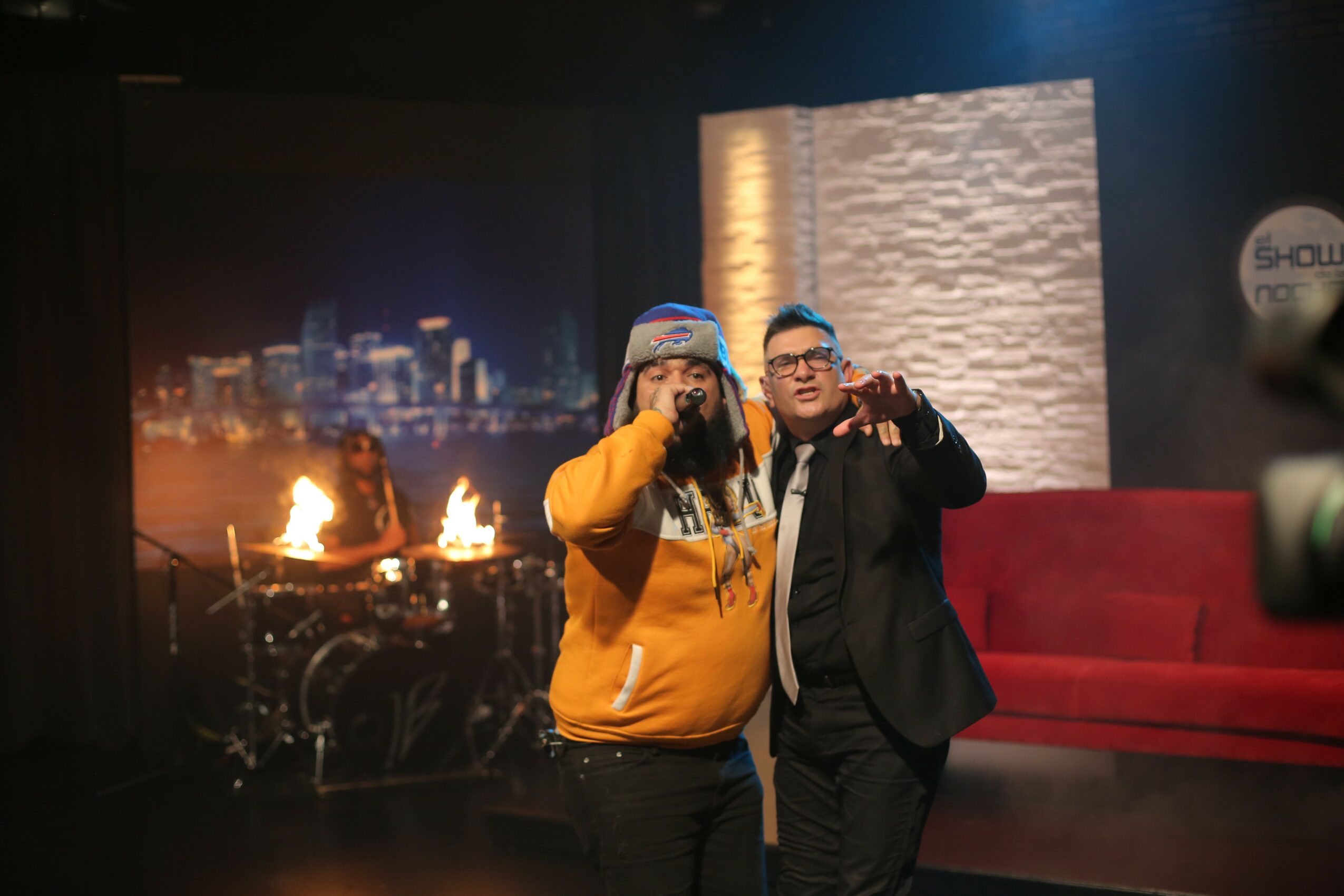 Two men pose for a photo in a studio with cityscape background and red couch, one in a hoodie and the other in a suit.