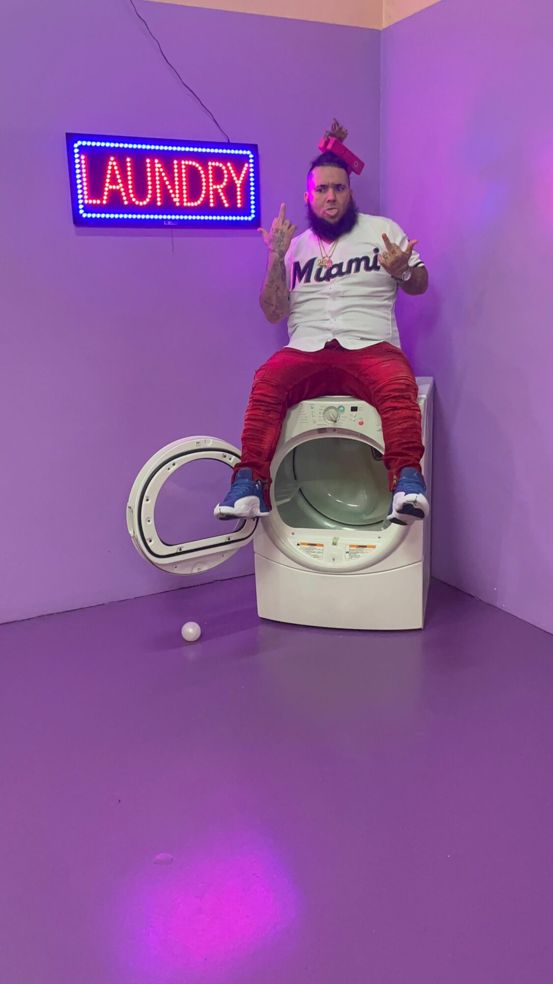 Person with pink hair and a headband stands next to a washing machine in a purple room, laundry sign on wall.