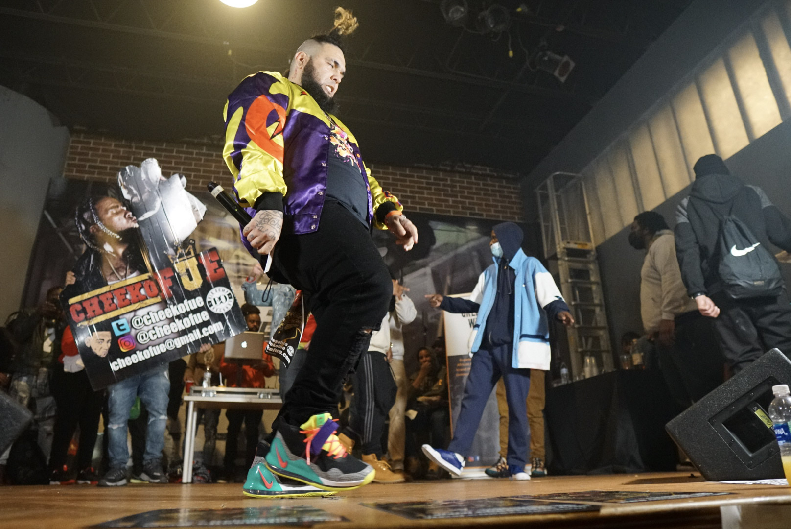Person dancing on stage with a crowd in the background, colorful clothing, and a poster in the scene.