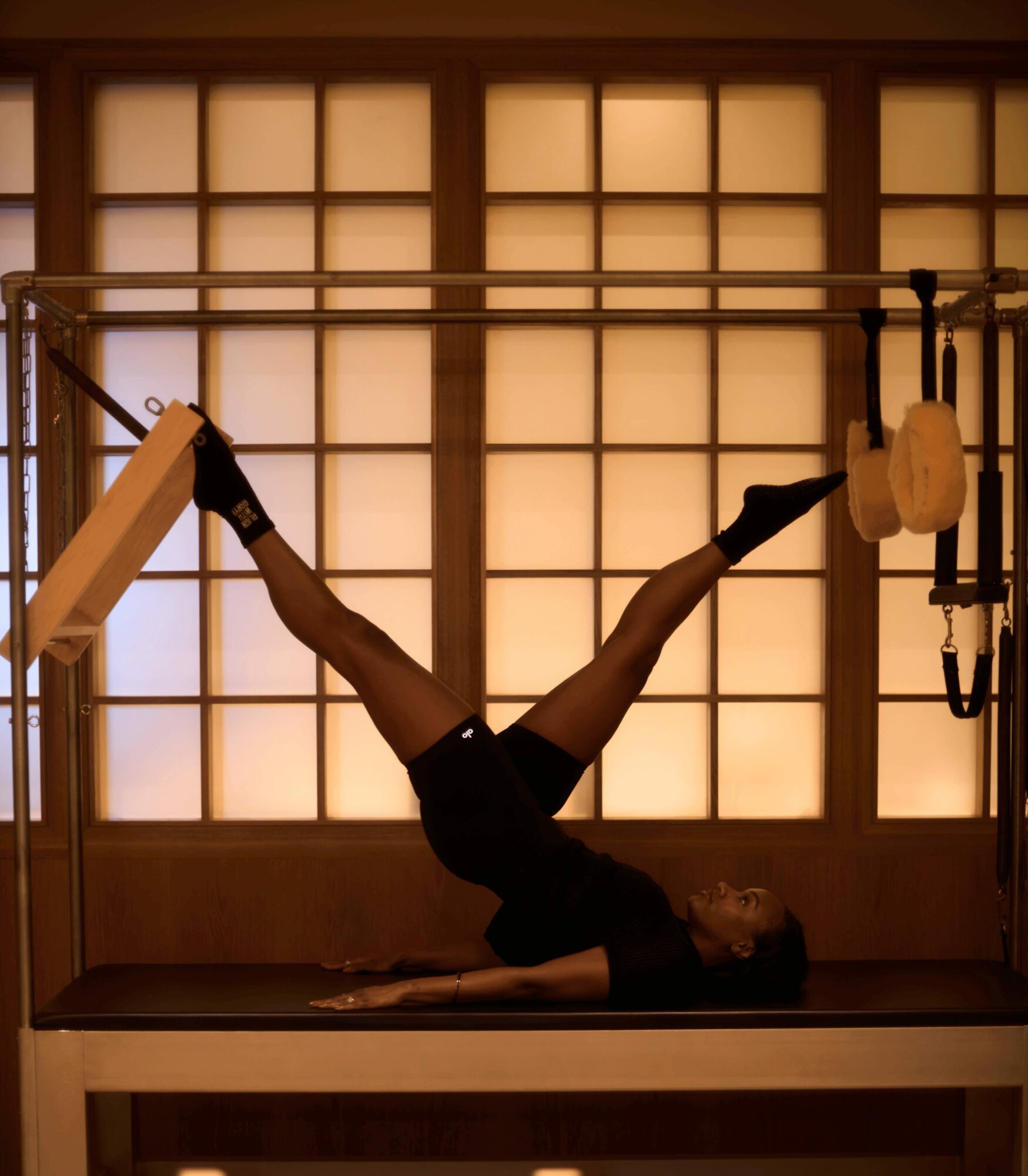 Person performing a split on a gymnastics or dance apparatus in a room with shoji-style sliding doors, wearing black clothing and gloves.