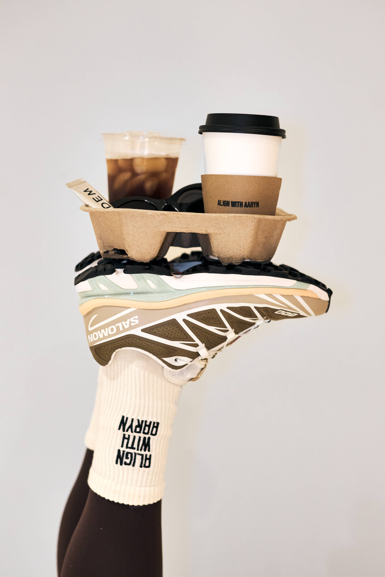 A hand wearing a white glove with black text holds a skateboard with two coffee cups on top, against a plain background.