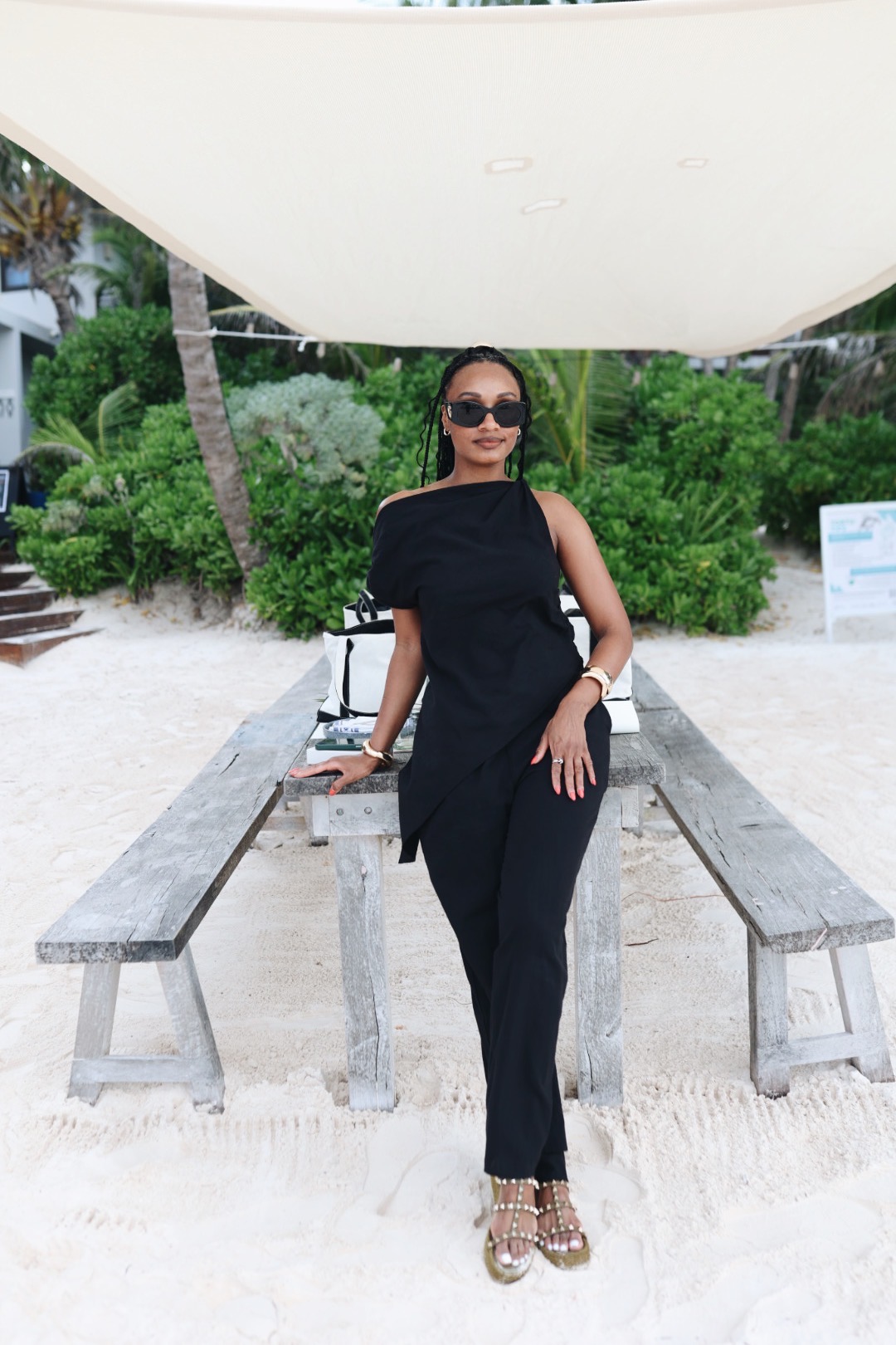 Woman in black outfit and sunglasses standing outdoors with greenery and benches behind her.