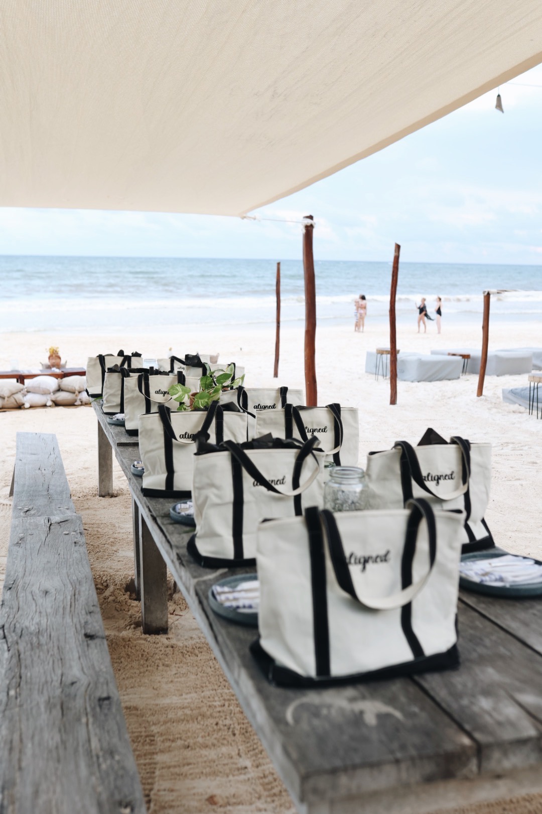 Beachside dining setup with bags and plates on a wooden table, ocean and people in the background, under a shaded area.