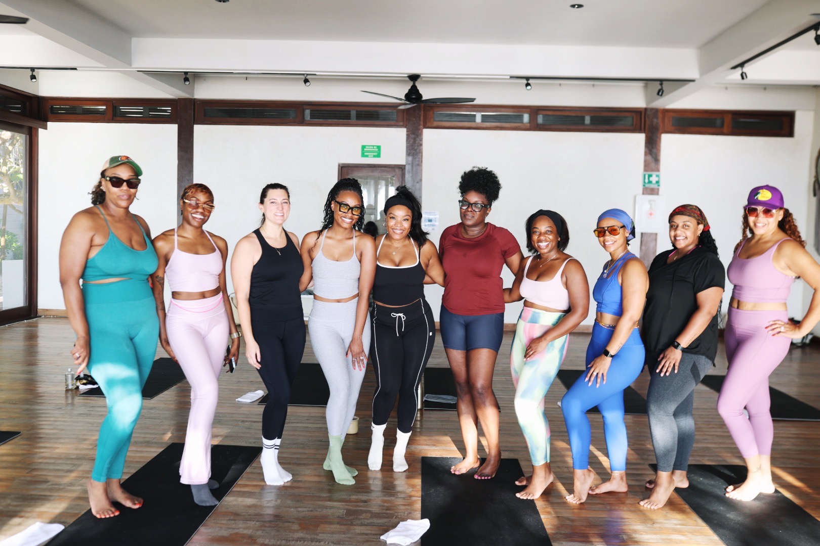 Group of ten women standing in a dance studio, smiling, with wooden floors and large windows behind them.