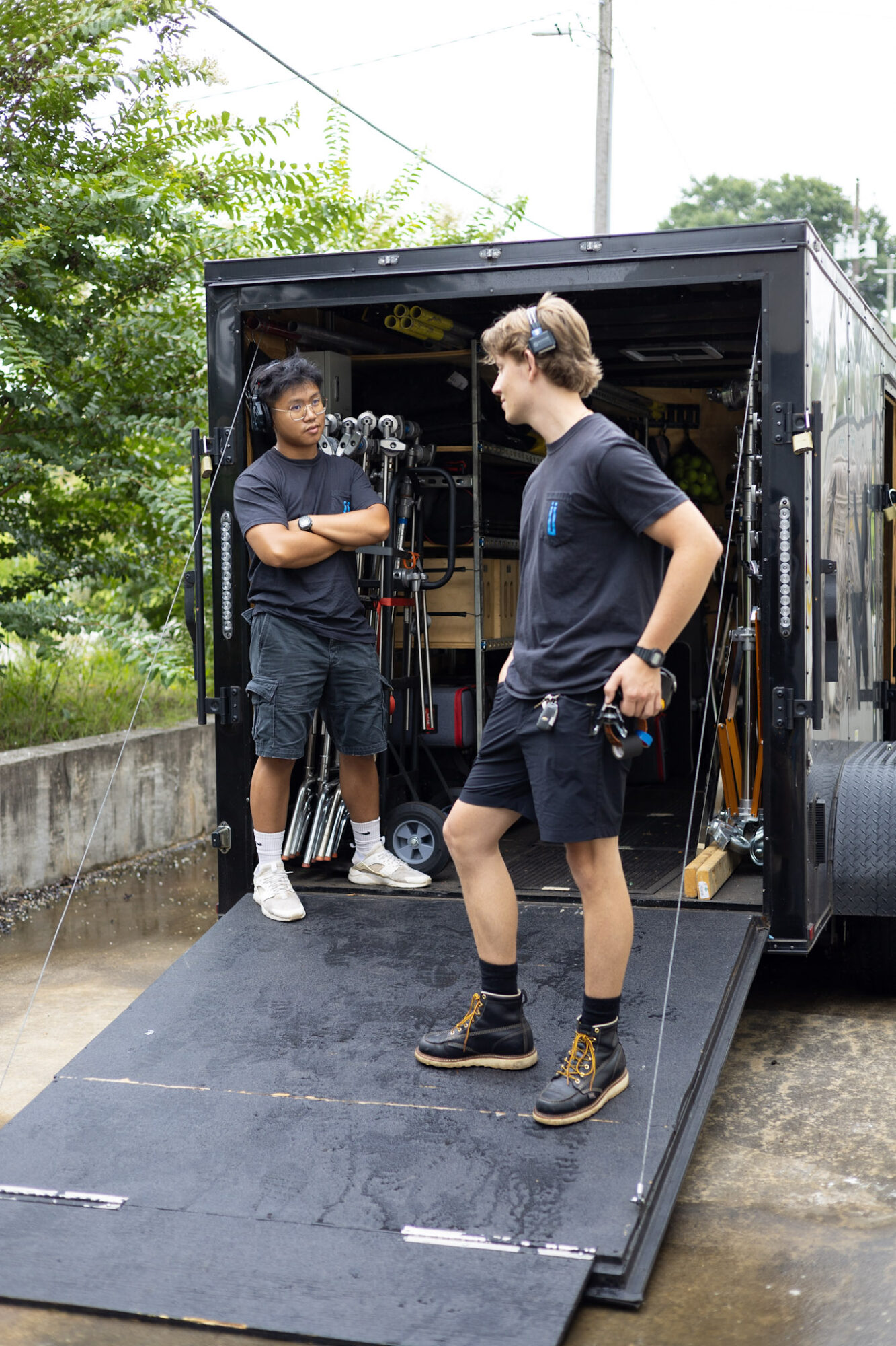 Two young men stand outside a black equipment trailer, one holding a camera and the other wearing gloves and boots.