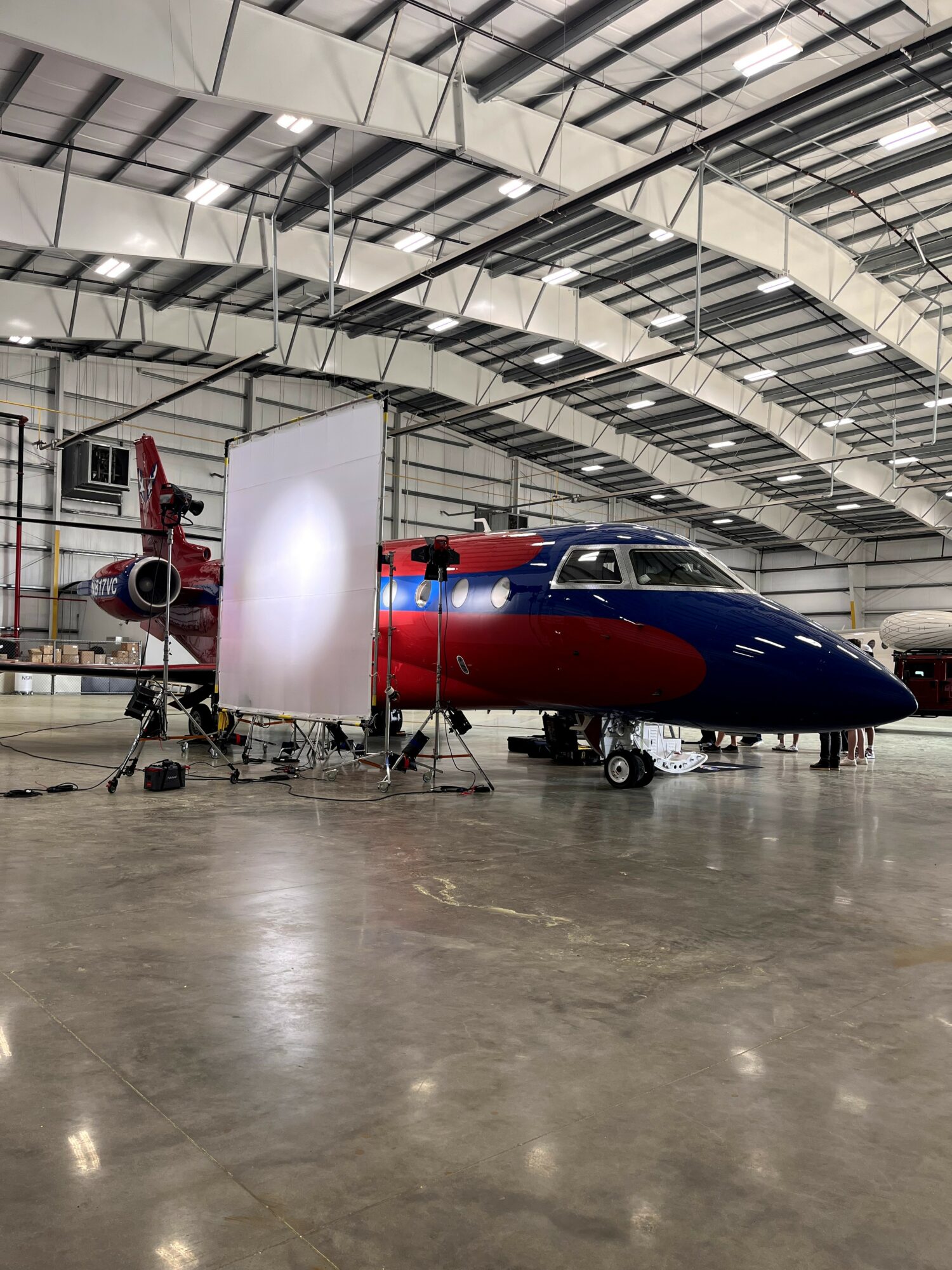 A red and blue aircraft inside a large hangar with a high ceiling and metal beams, surrounded by equipment and lighting.