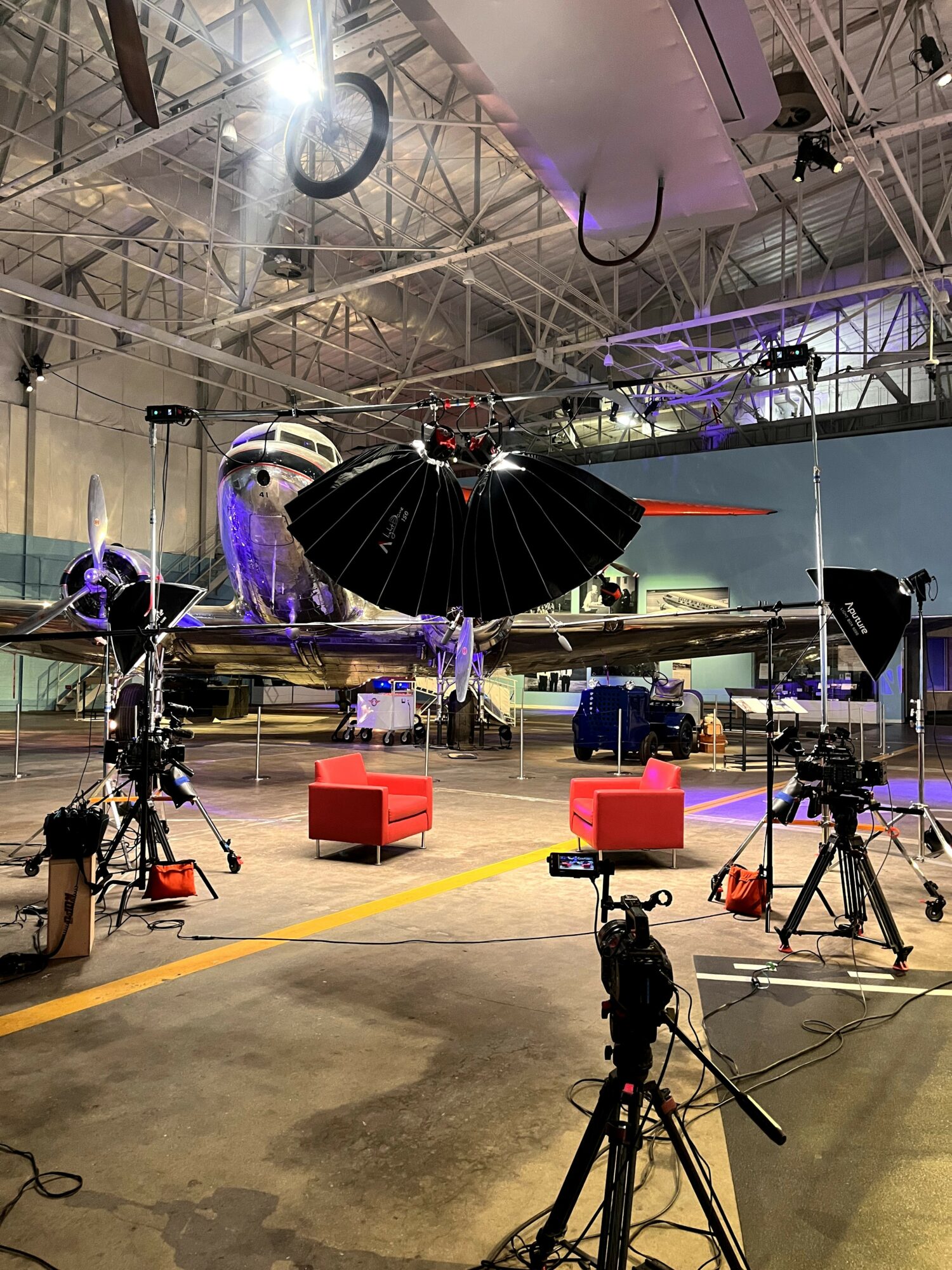Large drone with black umbrella-like propellers suspended in an industrial space with cameras and equipment, two red chairs below.