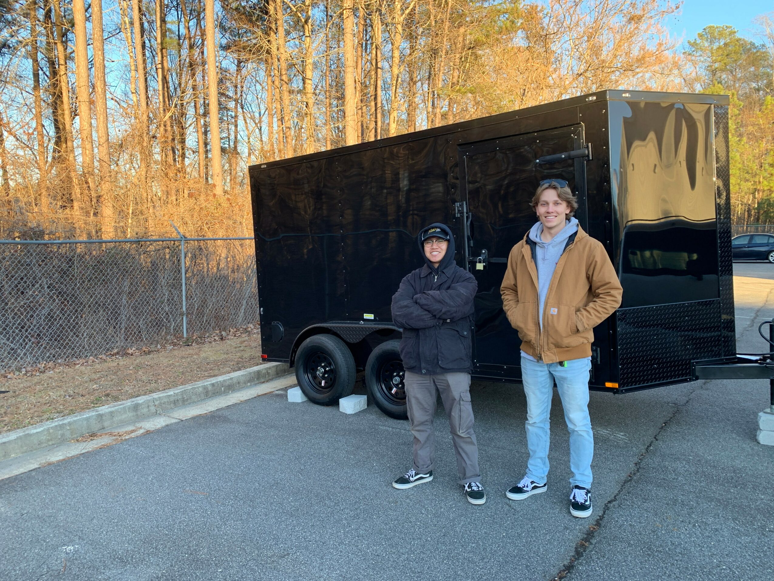 Two boys stand in front of a black trailer on a paved area with trees in the background.