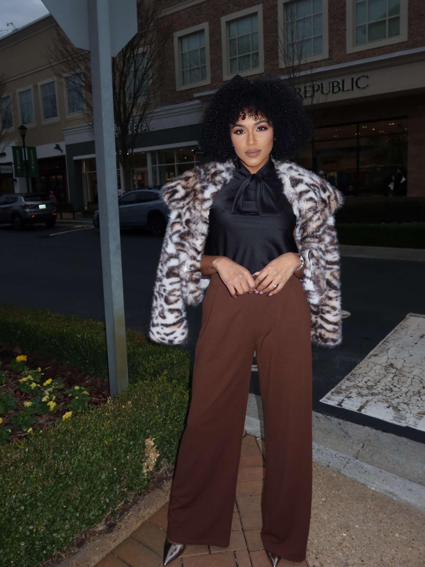 Woman with curly hair wearing a patterned coat, black blouse, and brown pants standing outdoors on a city sidewalk.